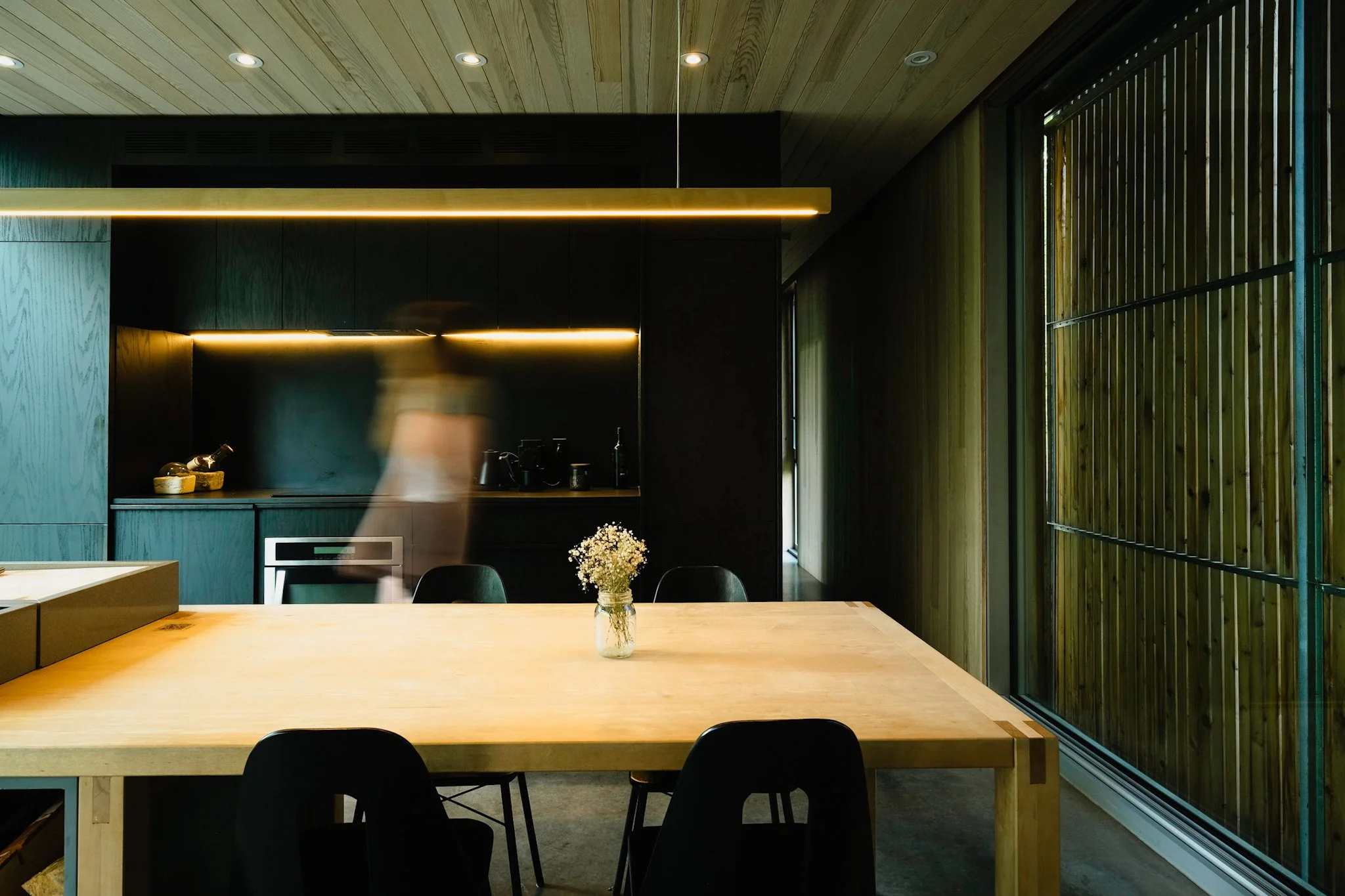 Modern dining area with a wooden table, black chairs, a small vase of flowers, dark kitchen cabinets, and large windows with wooden blinds.