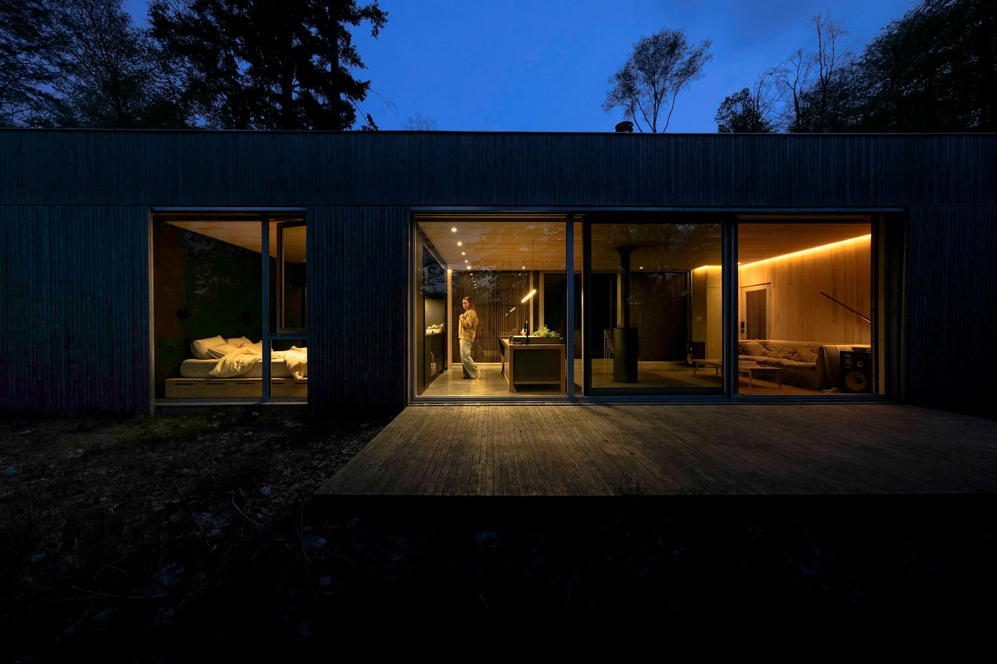 Modern house with large glass windows at dusk, showing an interior with a person in the kitchen, a bedroom on the left, and a living area on the right.