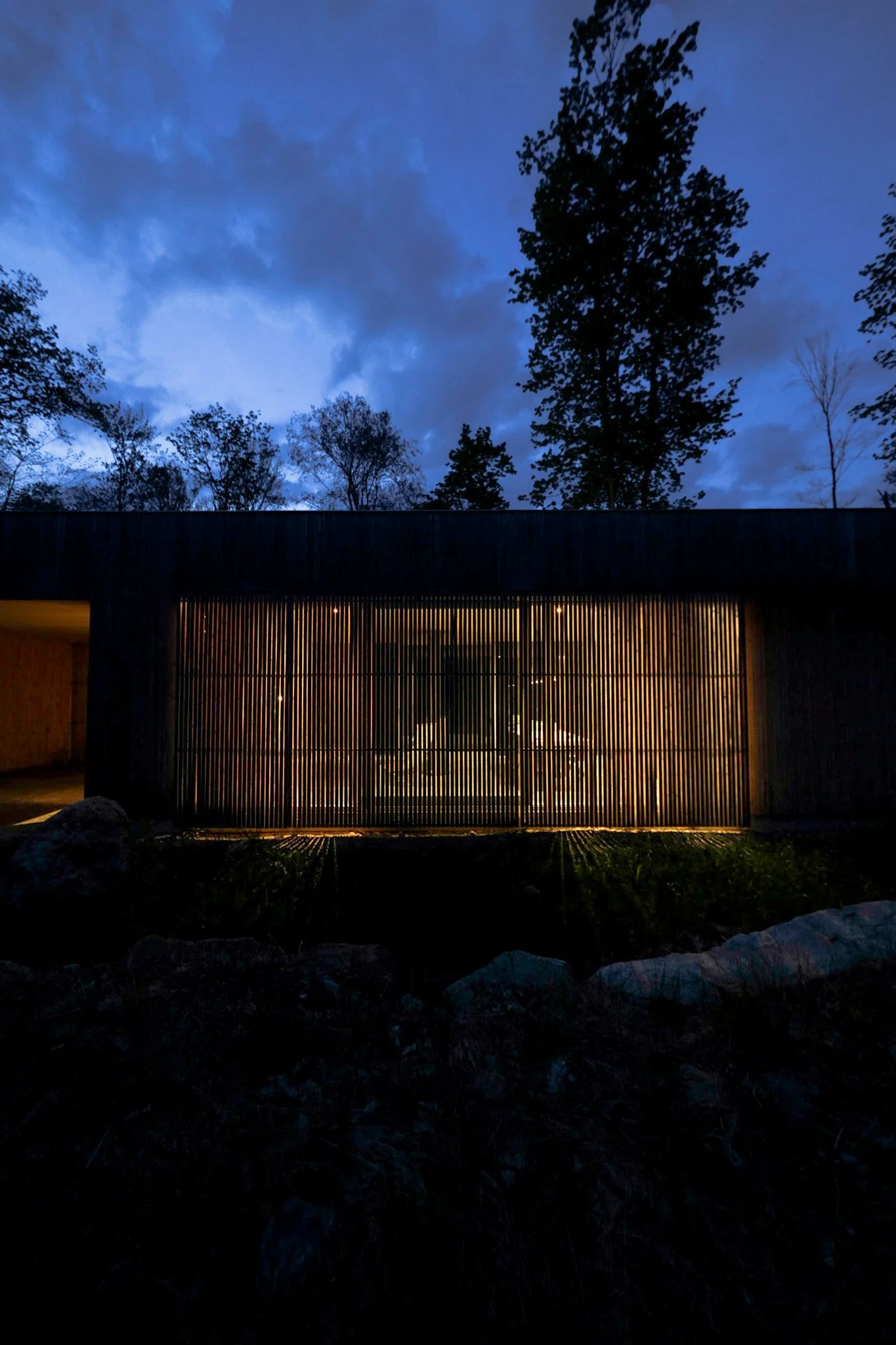 Modern building with wooden slat exterior and warm interior lighting, set against a dark blue evening sky with trees in the background.