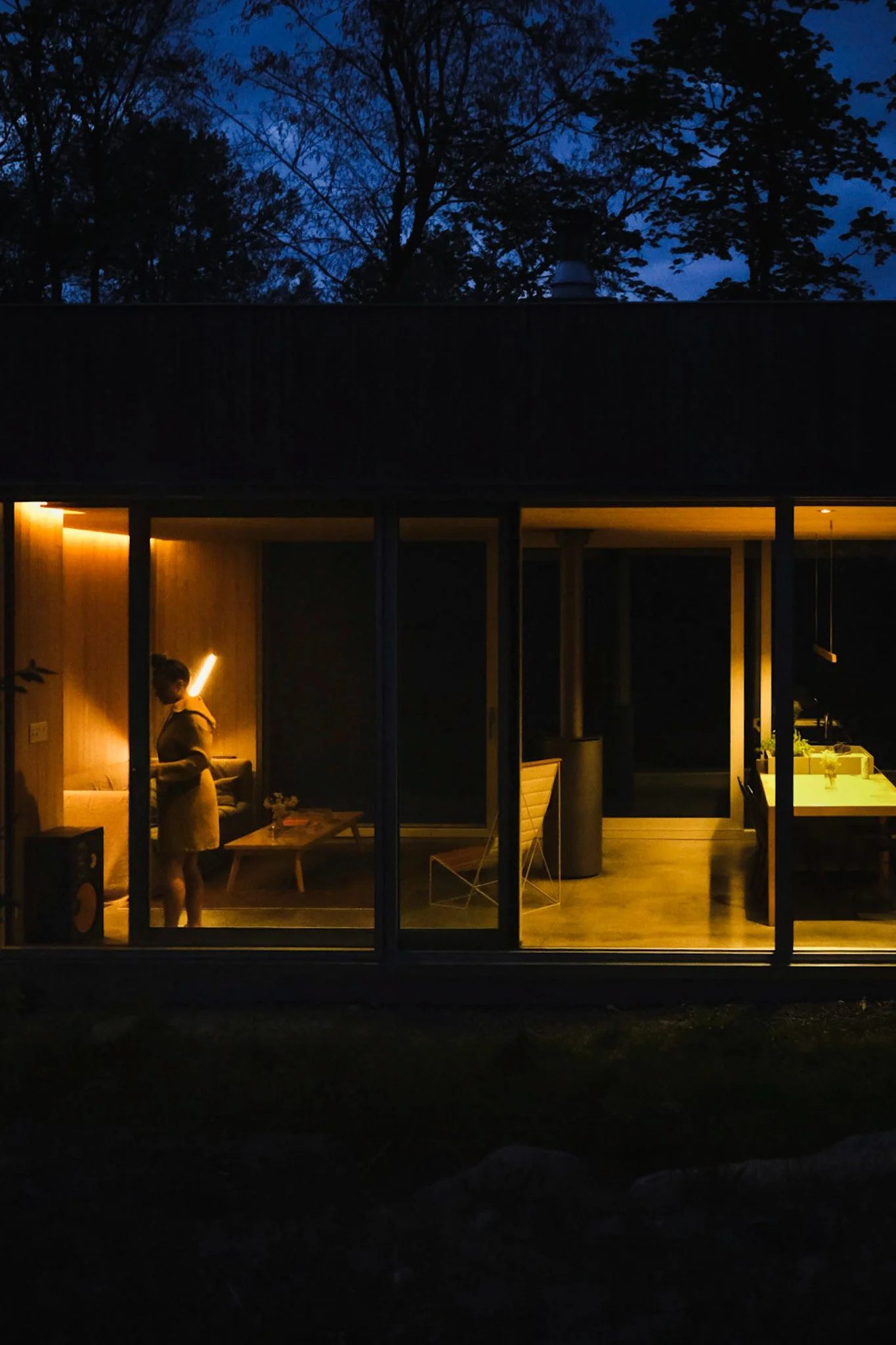 A woman stands inside a modern house with large glass windows, illuminated by warm yellow lighting, during twilight outside.