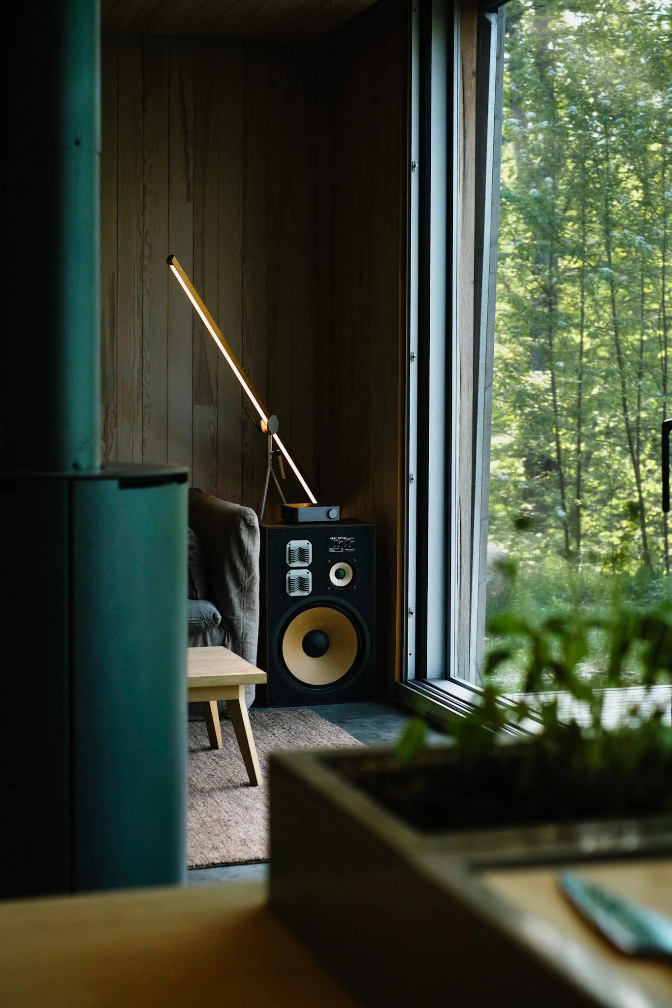 Interior of a modern living space with a large speaker near a floor-to-ceiling window showing green trees outside, with a small wooden table and a sofa.