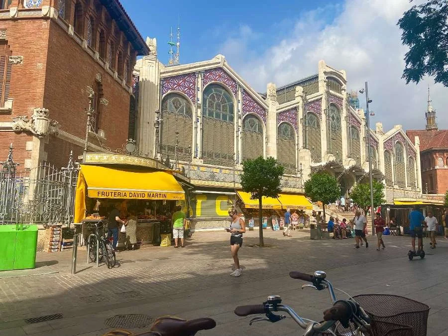 People stroll in front of a large building featuring bright yellow awnings at central market, Valencia.