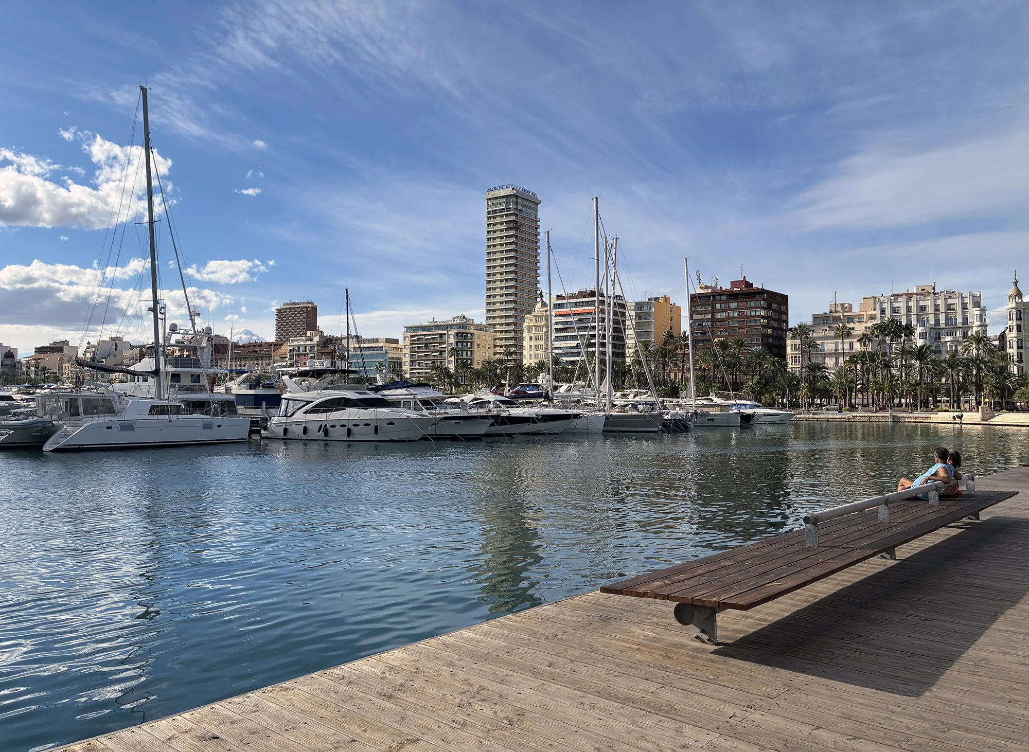 A couple on a bench in front of a marina in Alicante, Spain, takes in the peaceful atmosphere and scenic views.