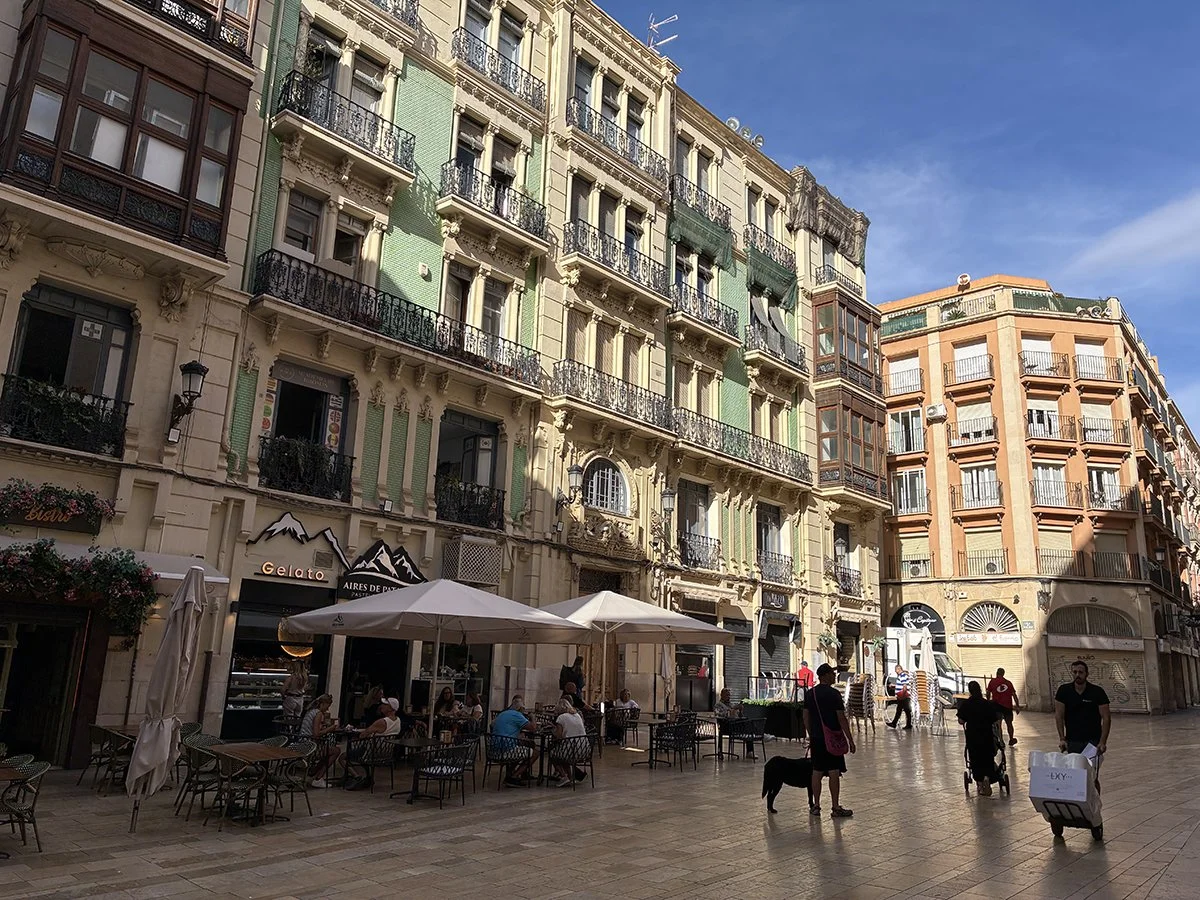 A peaceful Alicante city square features people walking among tables and umbrellas, showcasing a charming local vibe.