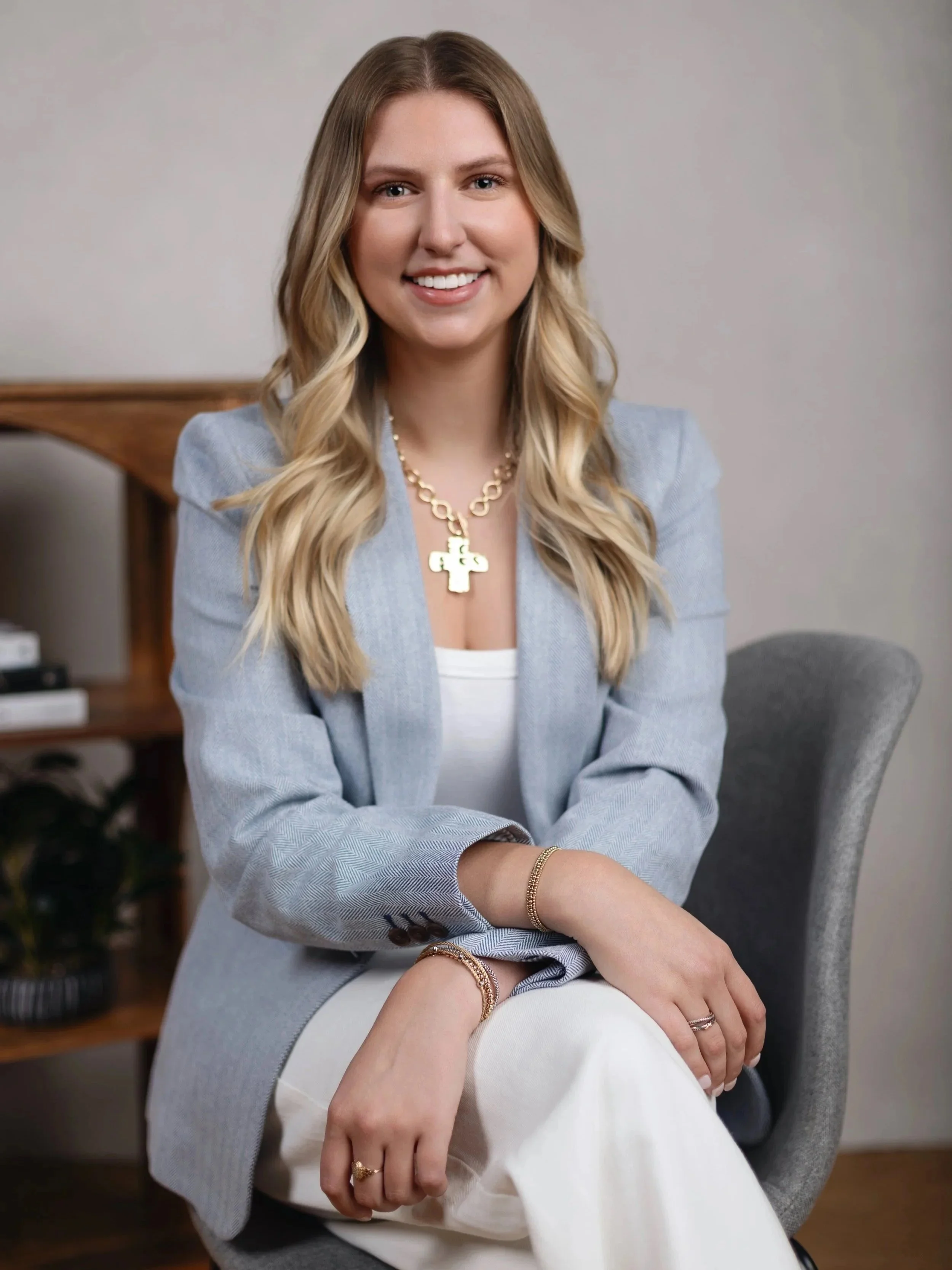 A young woman with long, wavy blonde hair smiling while sitting on a gray chair indoors. She is wearing a light gray blazer, white top, gold jewelry including a cross necklace and bracelets, and has multiple rings on her fingers. There is a wooden bookshelf with books and plants in the background.