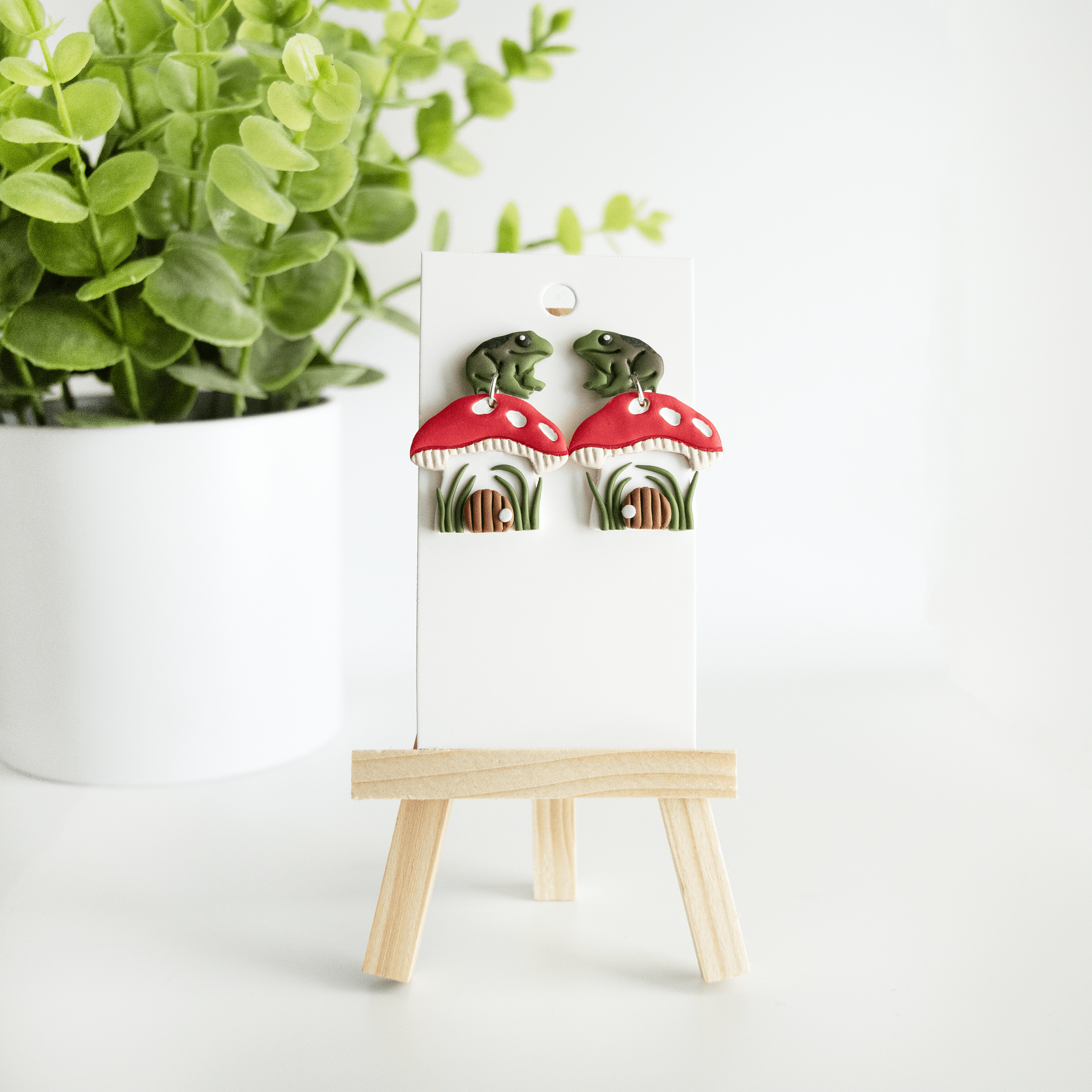 Photo of a pair of earrings with toads or frogs on top of a amanita mushroom house with a door and greenery