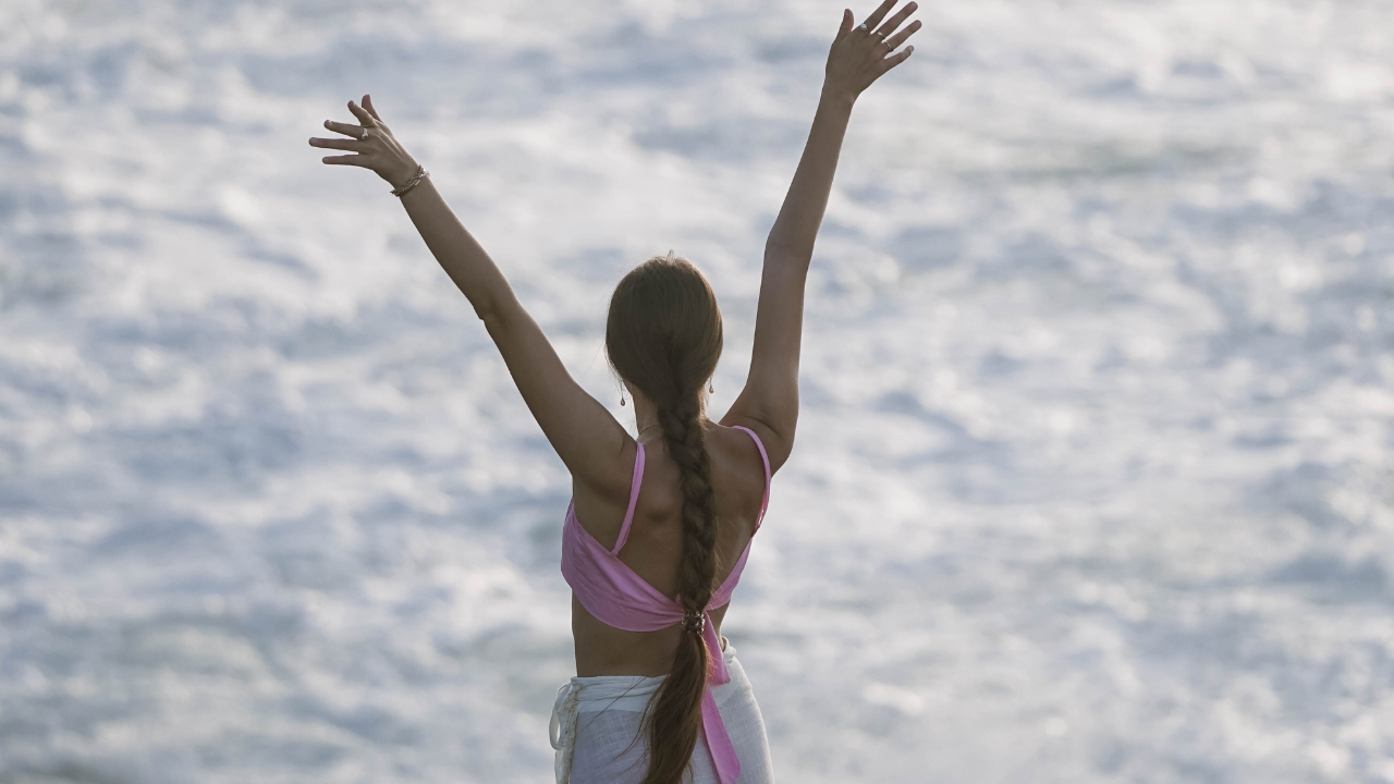 A woman with a braid, wearing a pink top and white skirt, stands on the beach with her arms raised toward the sky, facing the ocean.