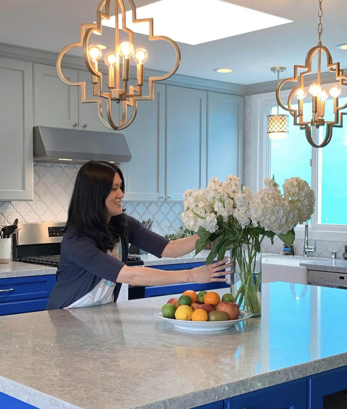 Woman arranging a bouquet of white hydrangeas on a kitchen island with a bowl of fruit nearby, modern kitchen with blue cabinets and decorative pendant lights.