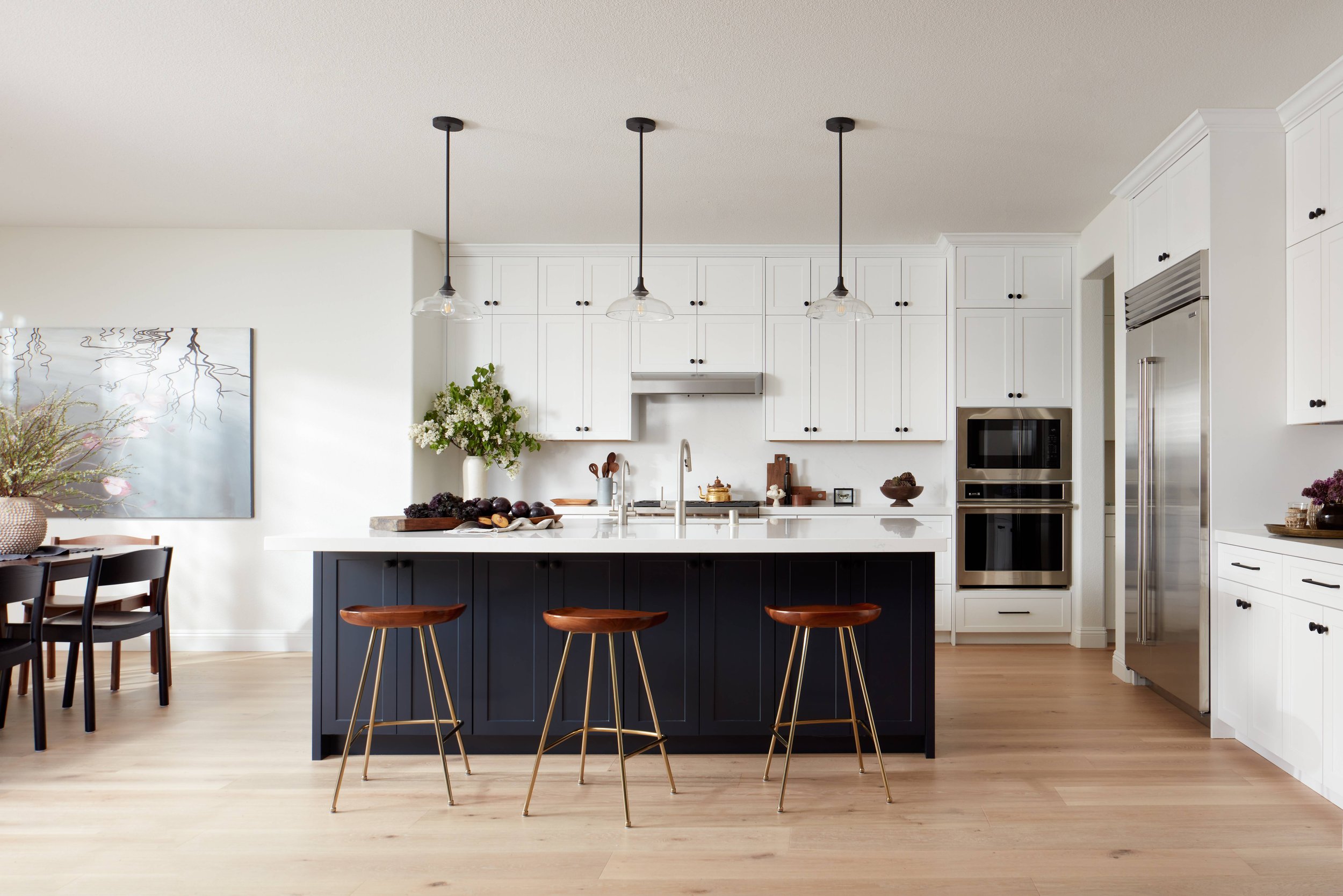 large kitchen with custom white cabinets, custom navy blue island, glass dome shaped light pendants, black hardware, black finishes, decor, artwork, walnut kitchen stools with brass legs, high-end appliances, quartz stone backsplash and countertop