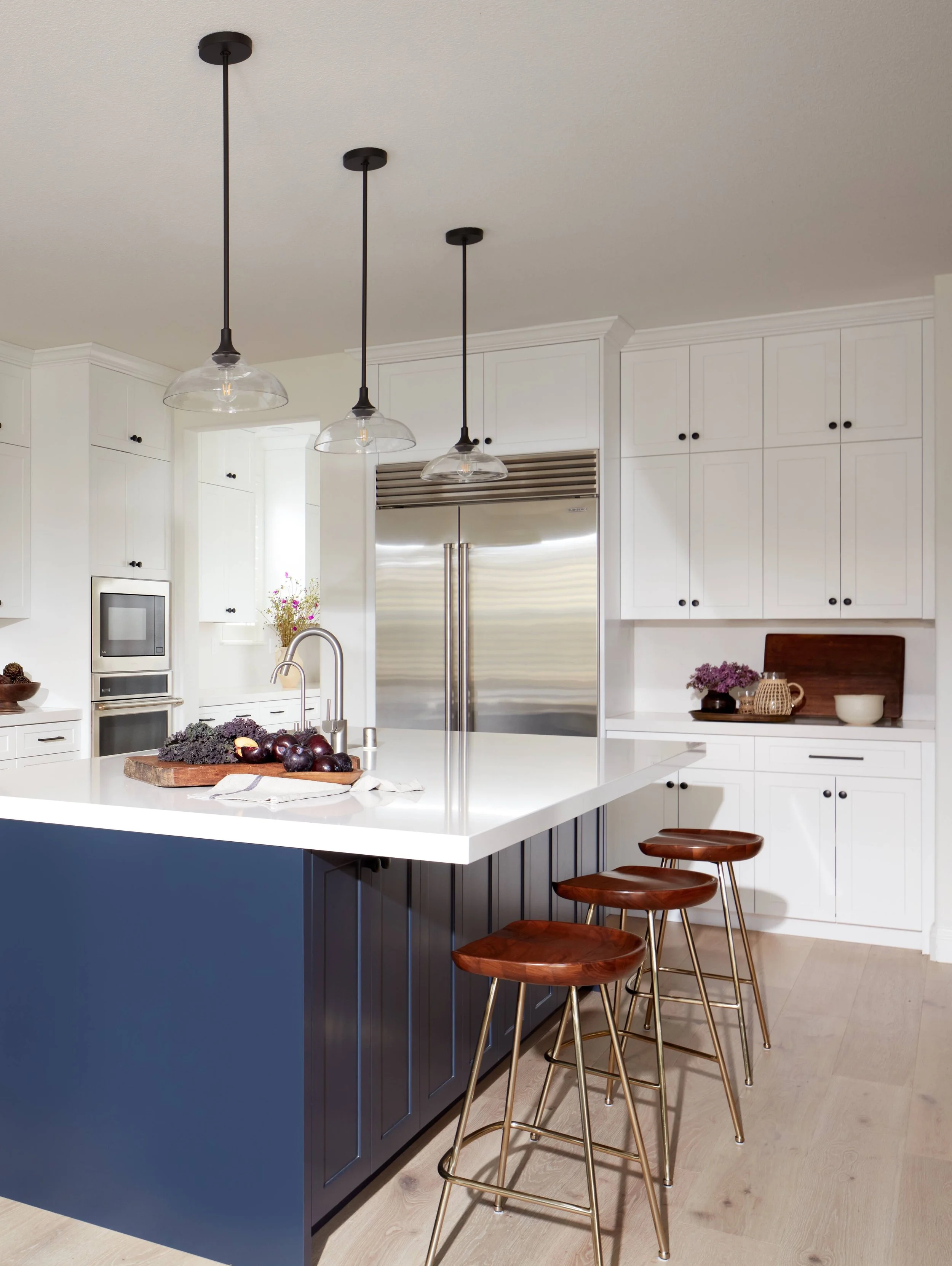 Modern kitchen with white cabinets, stainless steel refrigerator, and a kitchen island with a white countertop. Three pendant lights hang above the island, and several wooden barstools are lined up along one side of the island. There are purple flowers on the counter and a bowl of dark purple grapes on a cutting board.