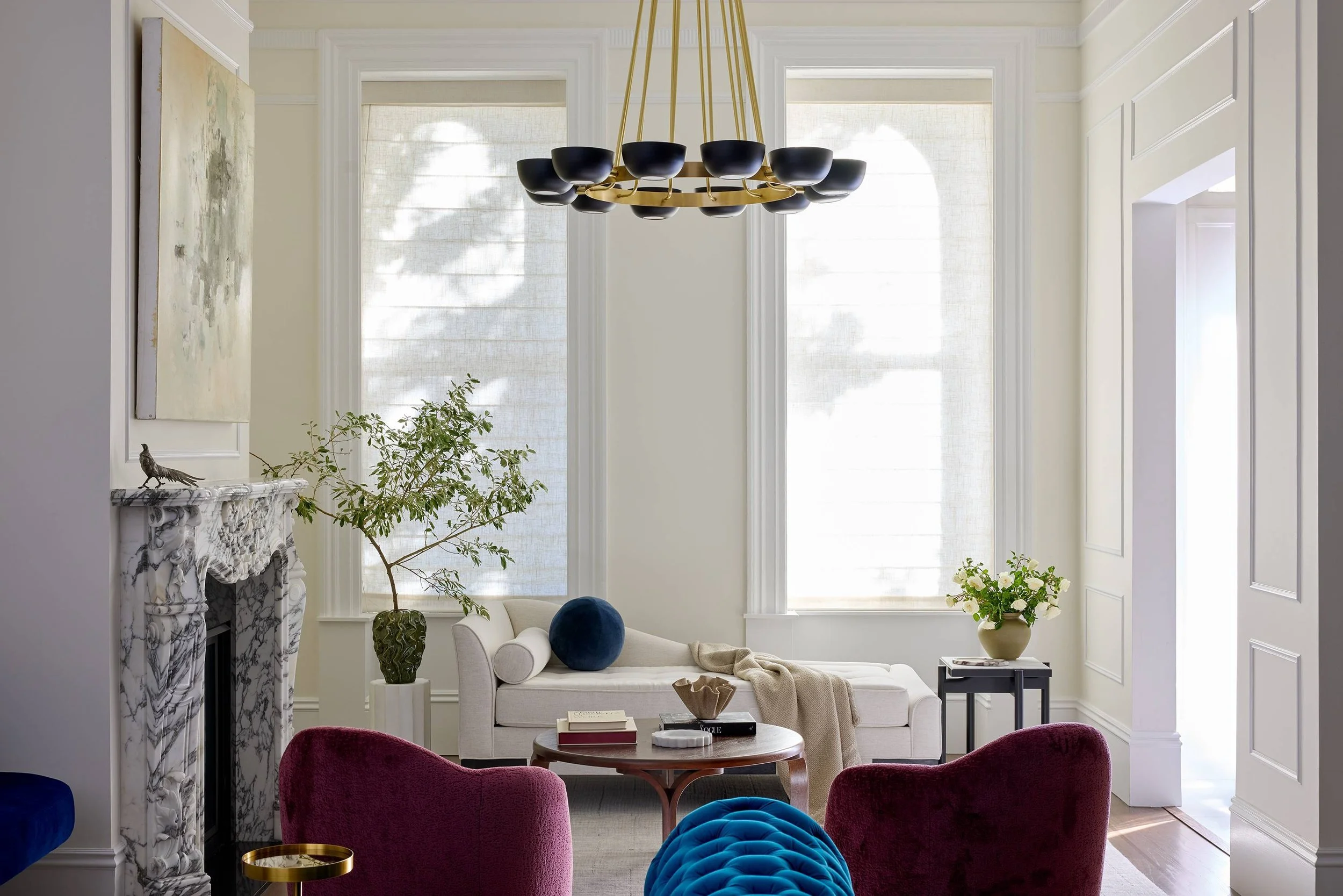 A living room with large windows, a white sofa, colorful velvet chairs, a marble fireplace, plants, and a modern chandelier.