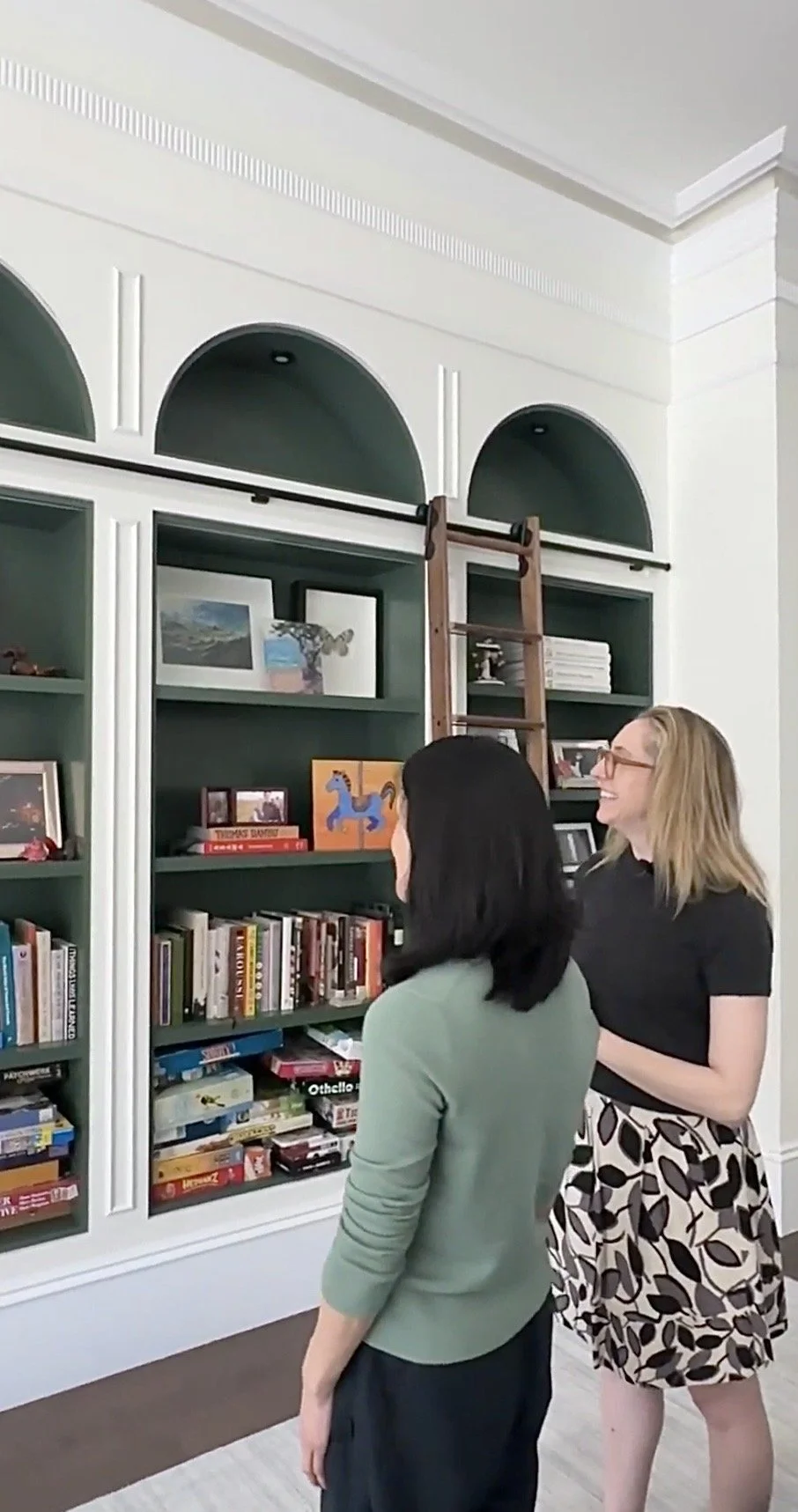 Principal designer, Alice and client admiring the 14 foot custom bookcase in green with arches, custom lighting, architectural details, molding, and walnut ladder.