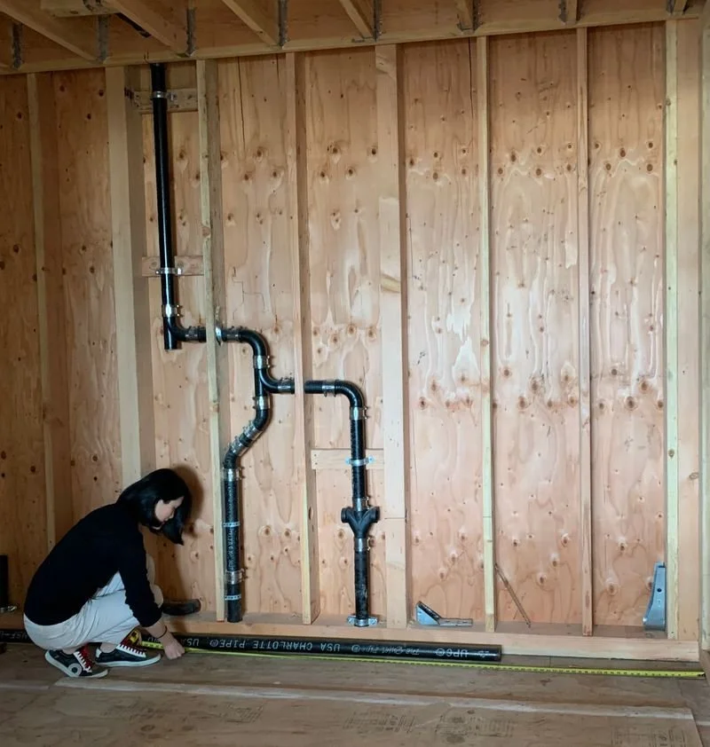 A person is measuring a black pipe system installed on a wooden wall of a building under construction.
