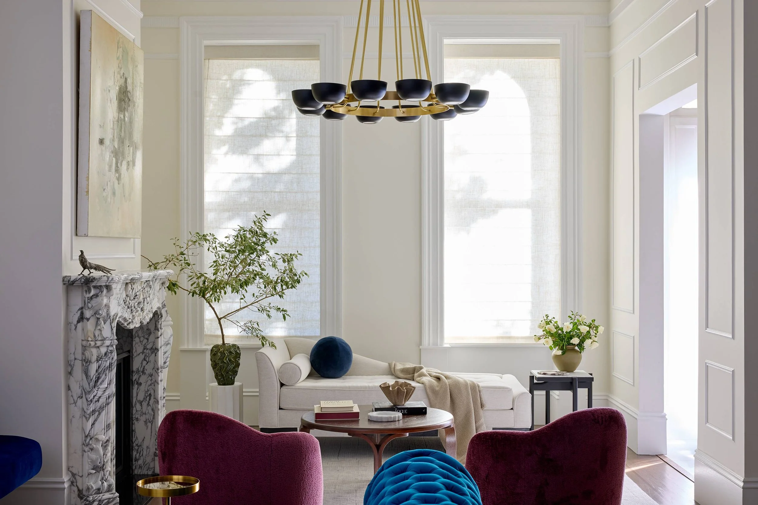 French Modern living room with modern black chandelier, velvet blue and wine furniture, artwork, large arched windows, white daybed chaise, and vintage wood cocktail table