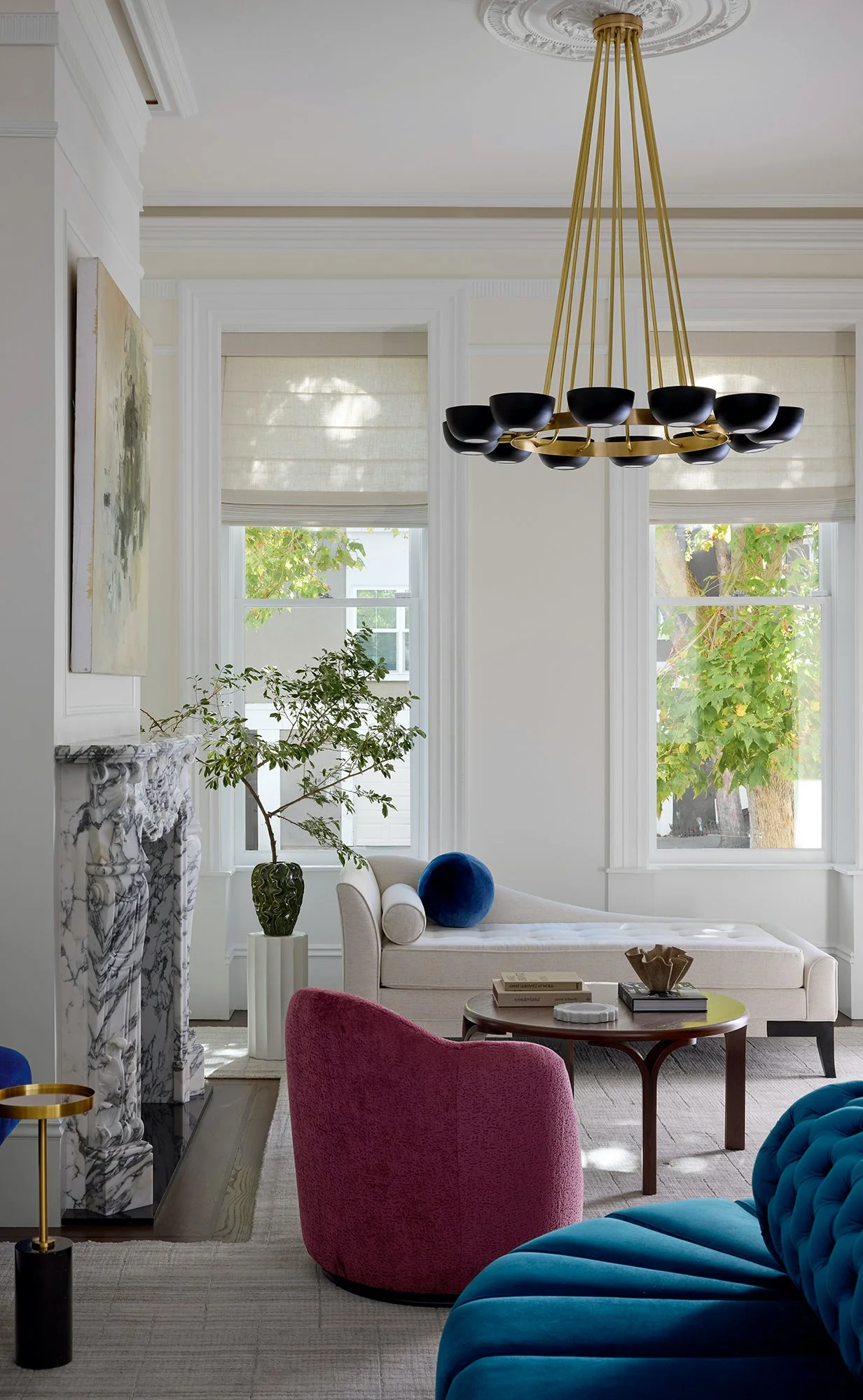 Grand Living room with white walls, architectural molding, marble fireplace, abstract painting, blue velvet sofa, wine velvet swivel chai, vintage round coffee table, tall window with natural light, large potted plant, sculptural black chandelier