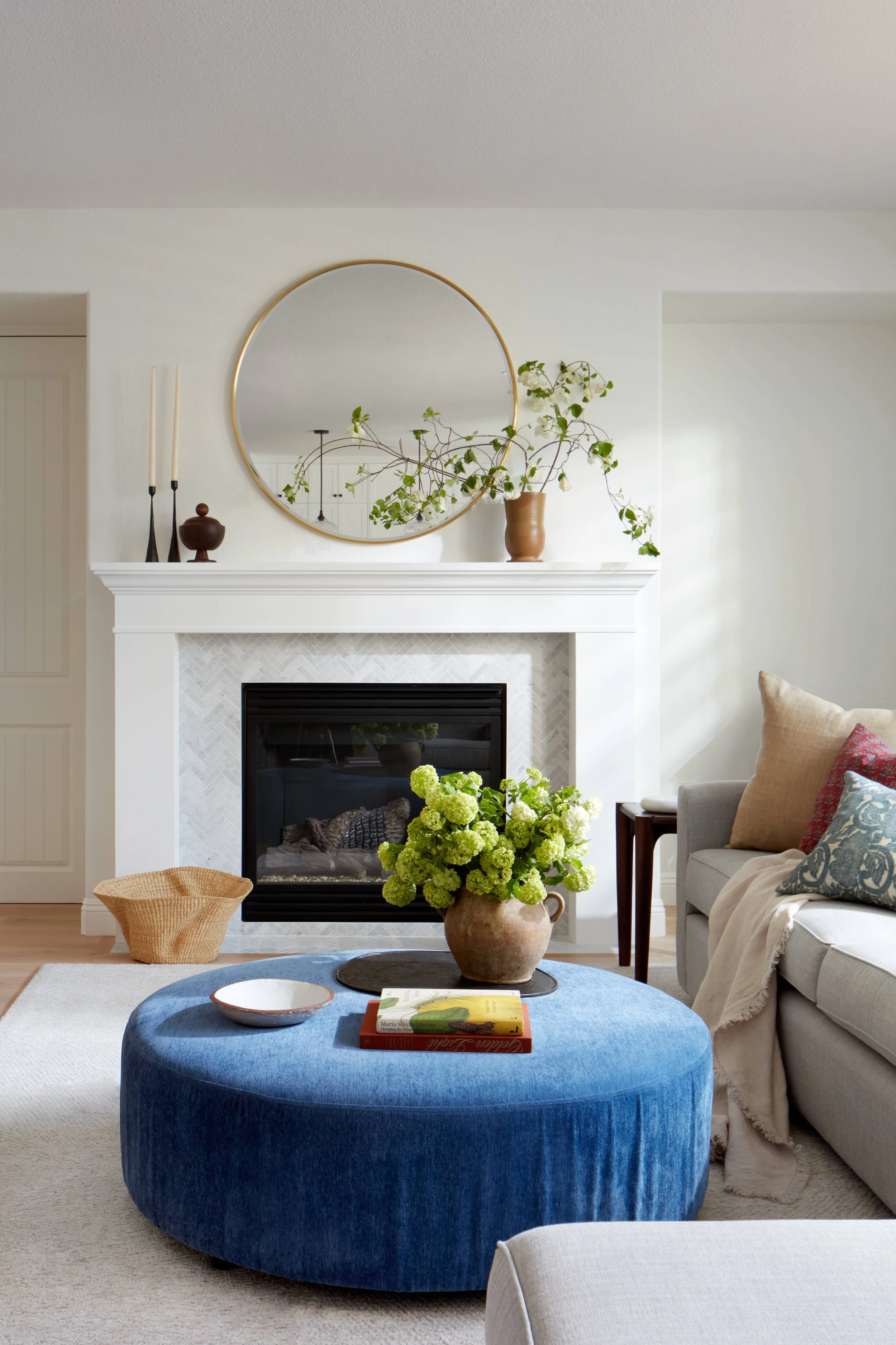 refined living room with white walls, gold accents, custom sofa sectional, custom round ottoman, round upholstered coffee table, neutral rug, engineered floors, gray herringbone marble tiles, custom fireplace