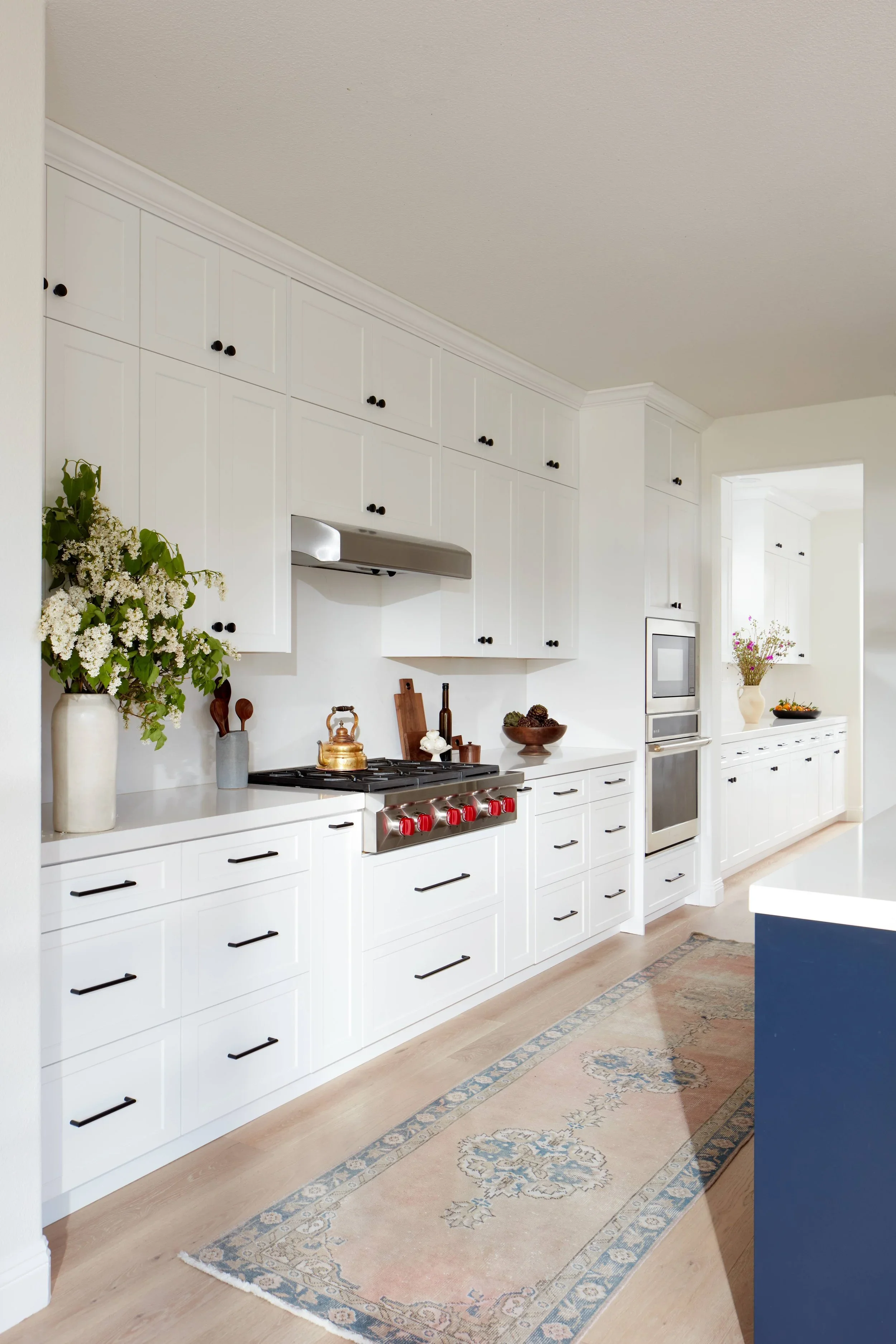 White kitchen with black knobs, stainless steel appliances, a beige rug, a light wood floor, green plants, and flowers in vases.