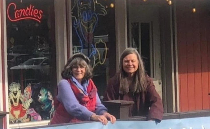 Two women smiling and leaning on a railing behind a large banner at WhaleFest Monterey 2025, scheduled for April 12-13, 2025, at Old Fisherman's Wharf, promoting inspiration, education, exploration, and empowerment.