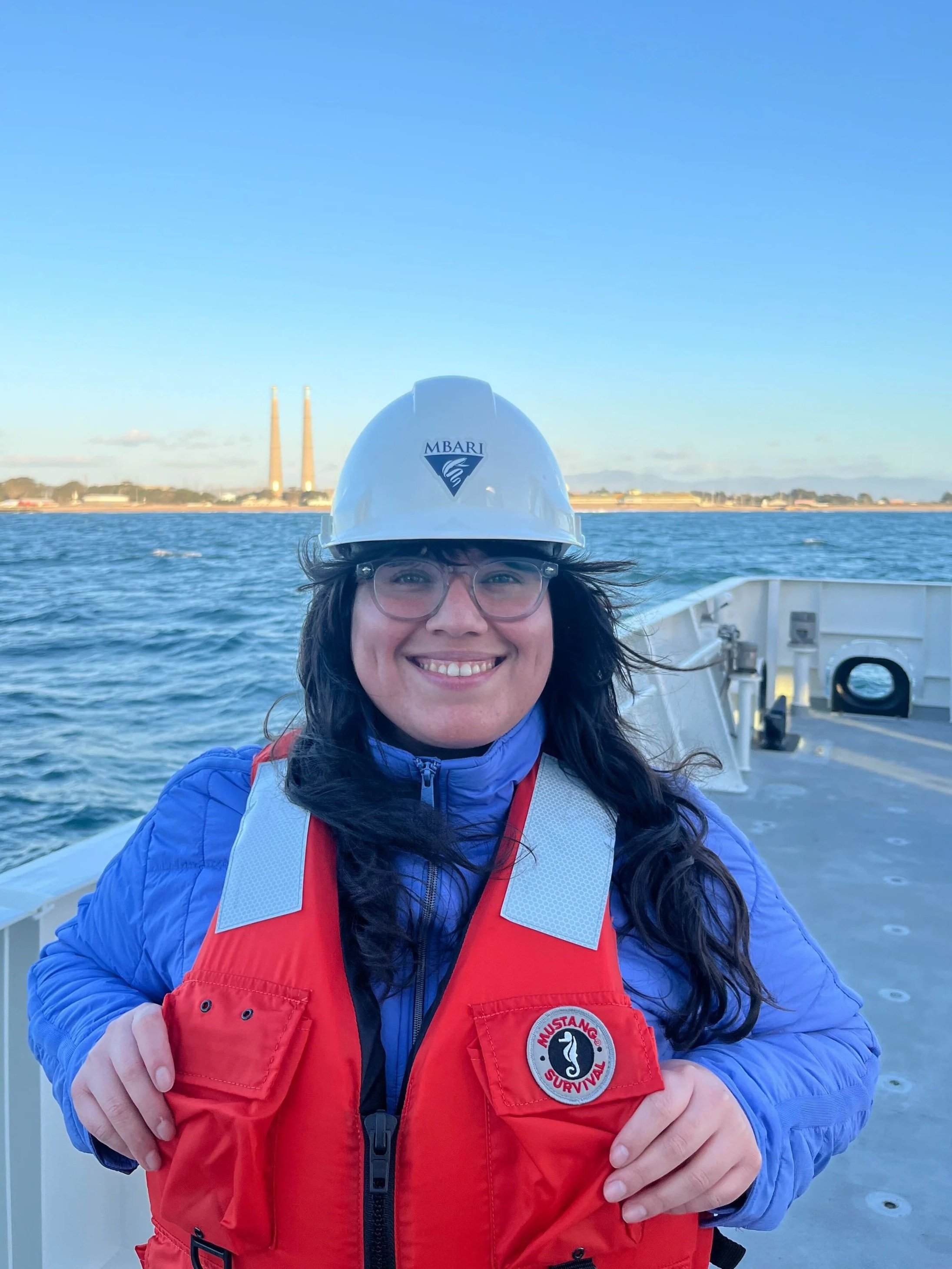 Photo of person with long black hair wearing a hard hat with a. long sleeve shirt with a bright color vest standing on a boat with water and land in the background with blue sky.