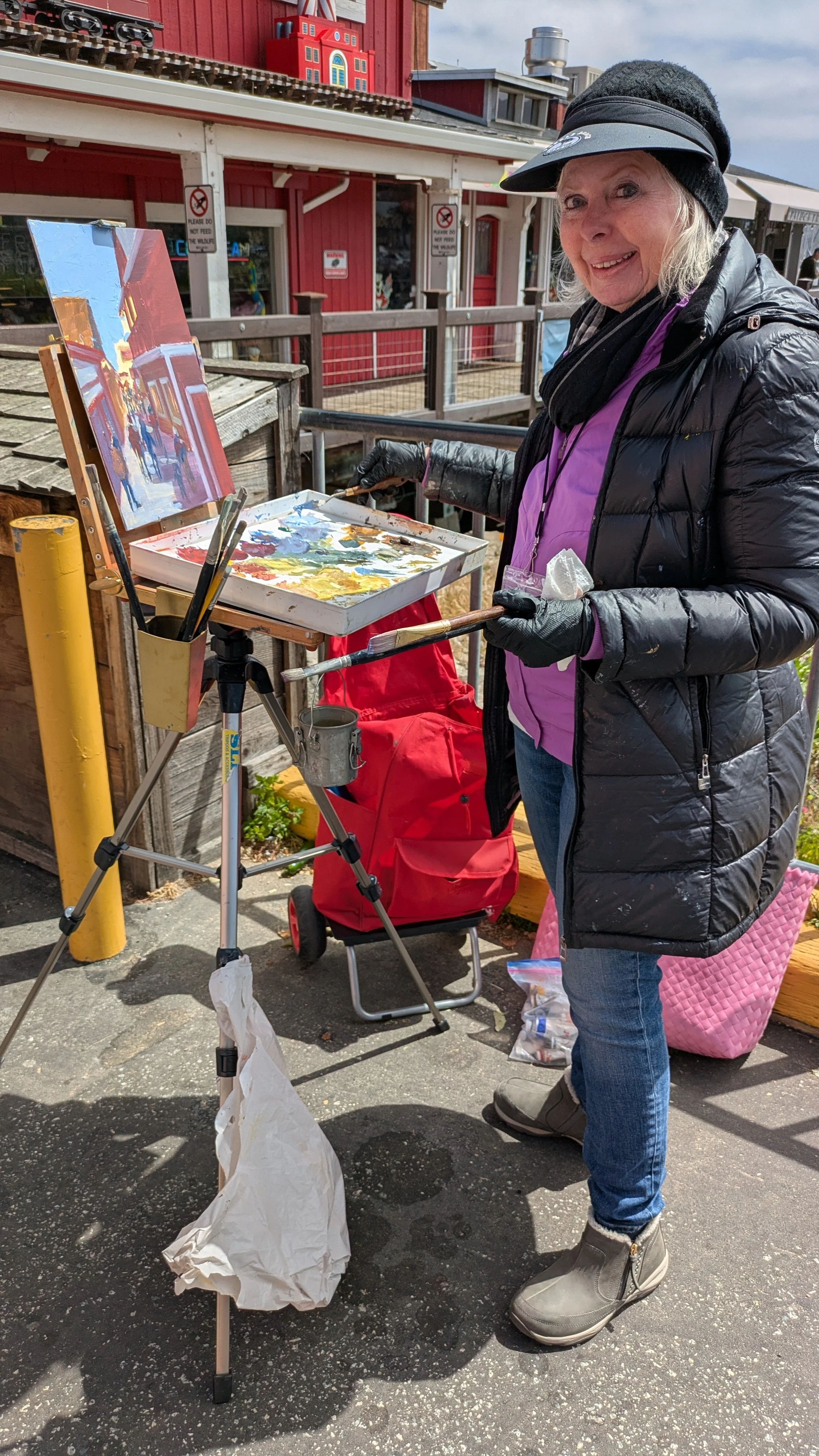 An artist standing near art easel with wharf in background.  Person is wearing a hat, jacket, pants, gloves.