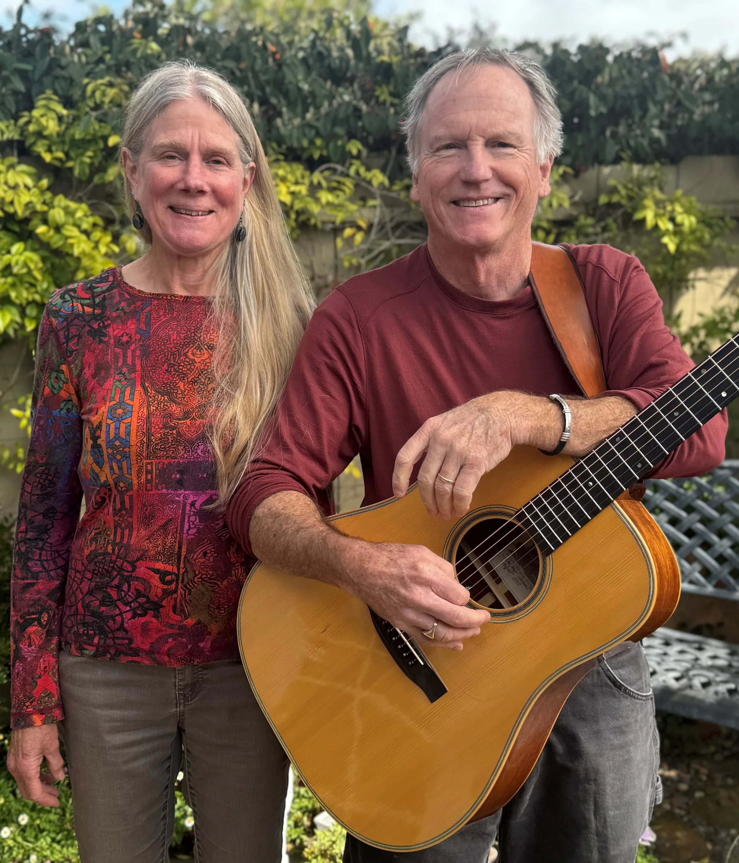 Two people standing looking at the camera, one with long gray hair, the other with short gray hair. Both wearing a burgundy color top.. the man is holding a guitar.