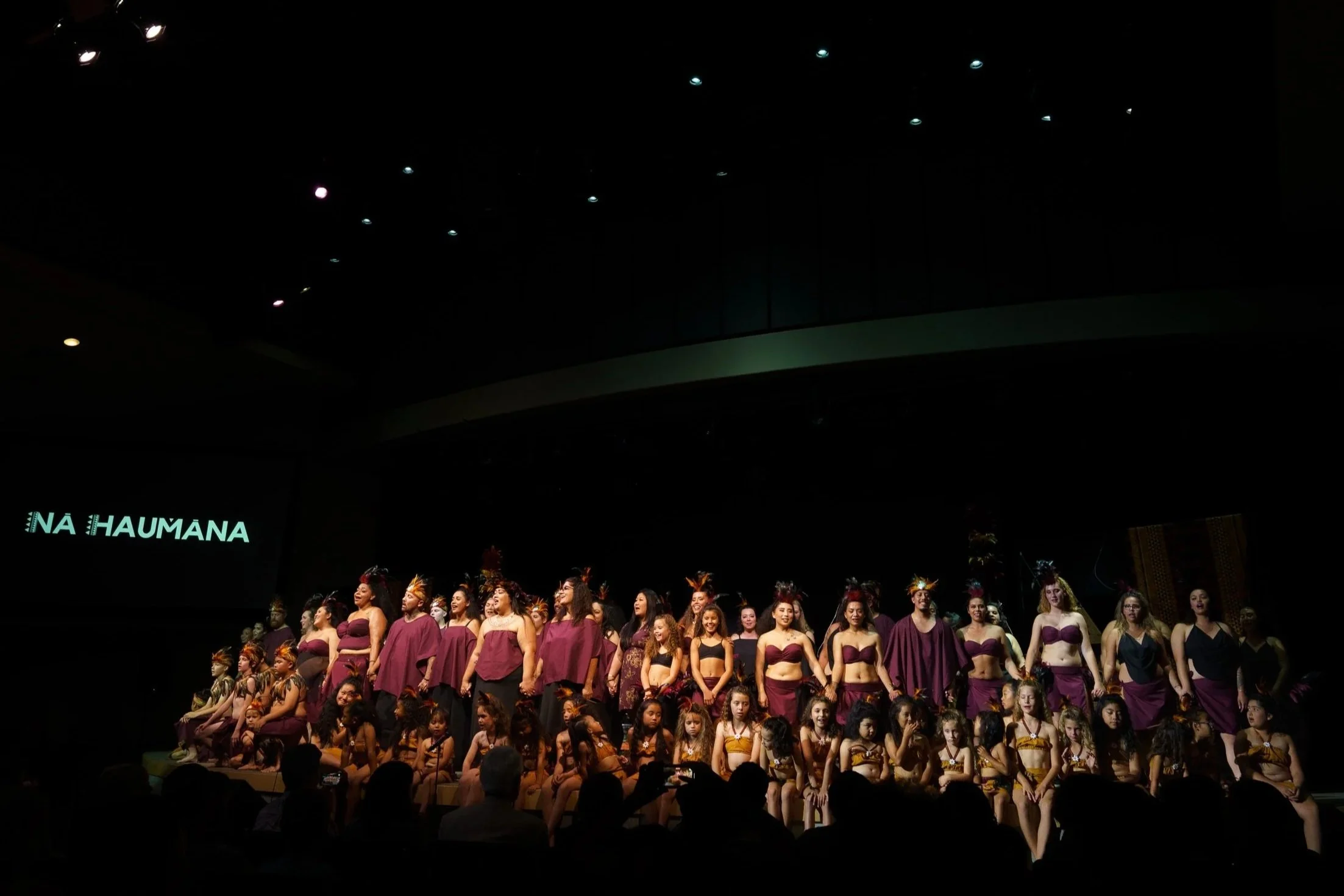 Large group of performers wearing burgundy color costumes standing on a large stage with a dark background and scene.