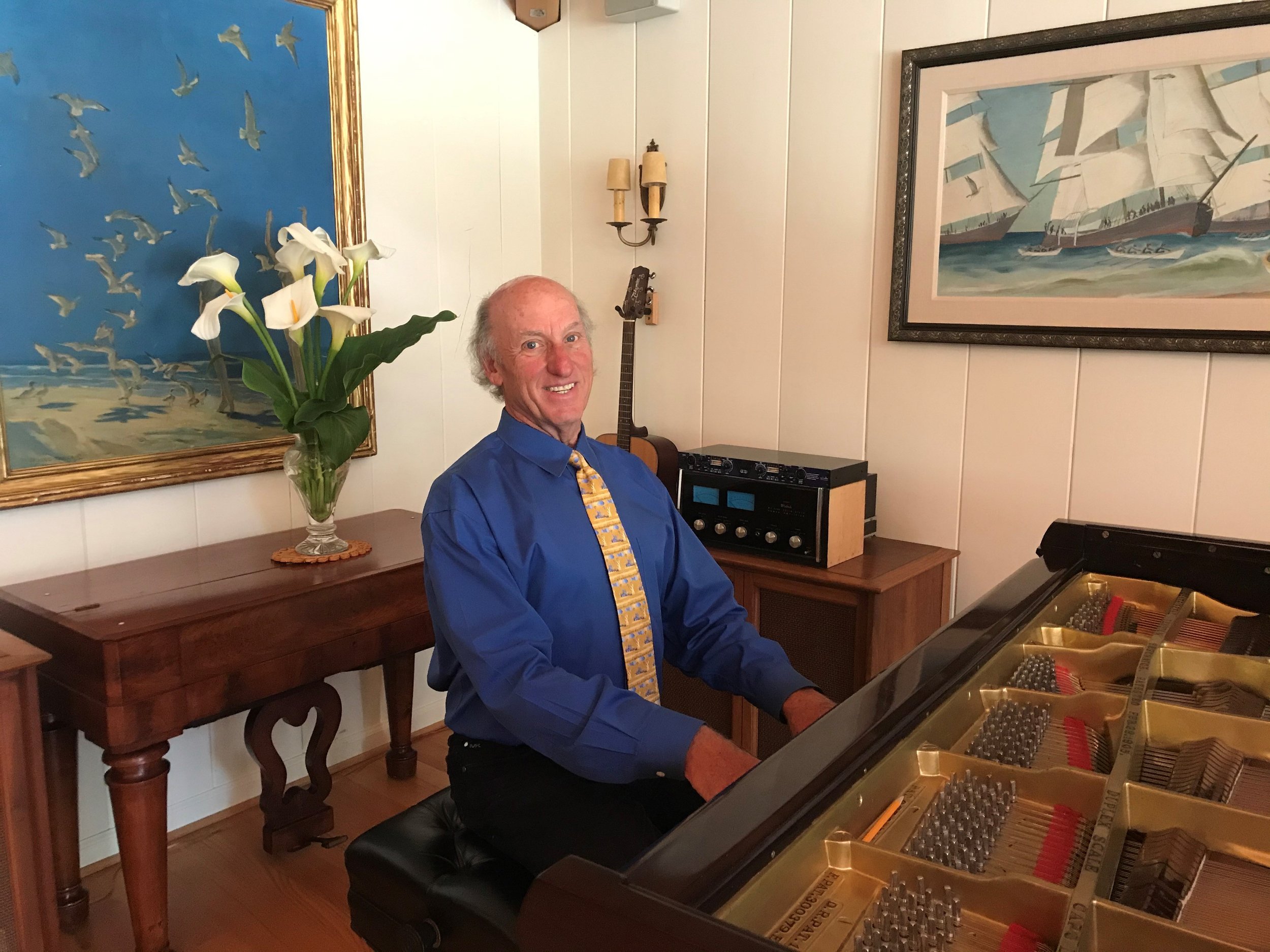Man sitting down playing a piano wearing a blue shirt and tie. with two portraits hanging on the wall behind him.