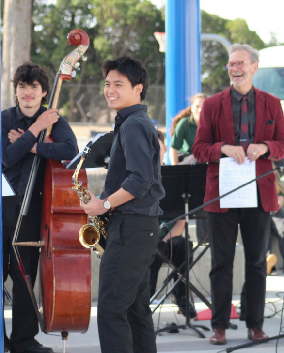 Three people smiling. Two in foreground, wearing black tops and one in background, wearing a black top with tie and burgundy jacket. Musical performance..