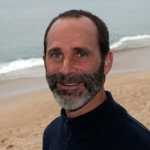 Person standing in front of beach and o. ce and ocean wave wearing a dirt color pop. with a salt and pepper beard and mustache