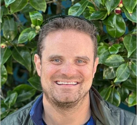 Person looking at the camera smiling with short, brown hair scruff on the face. standing in front of a greenbush.