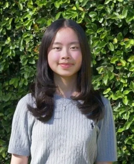 Photo of person standing in front of a greenbush, smiling with long brown hair wearing a sweater.