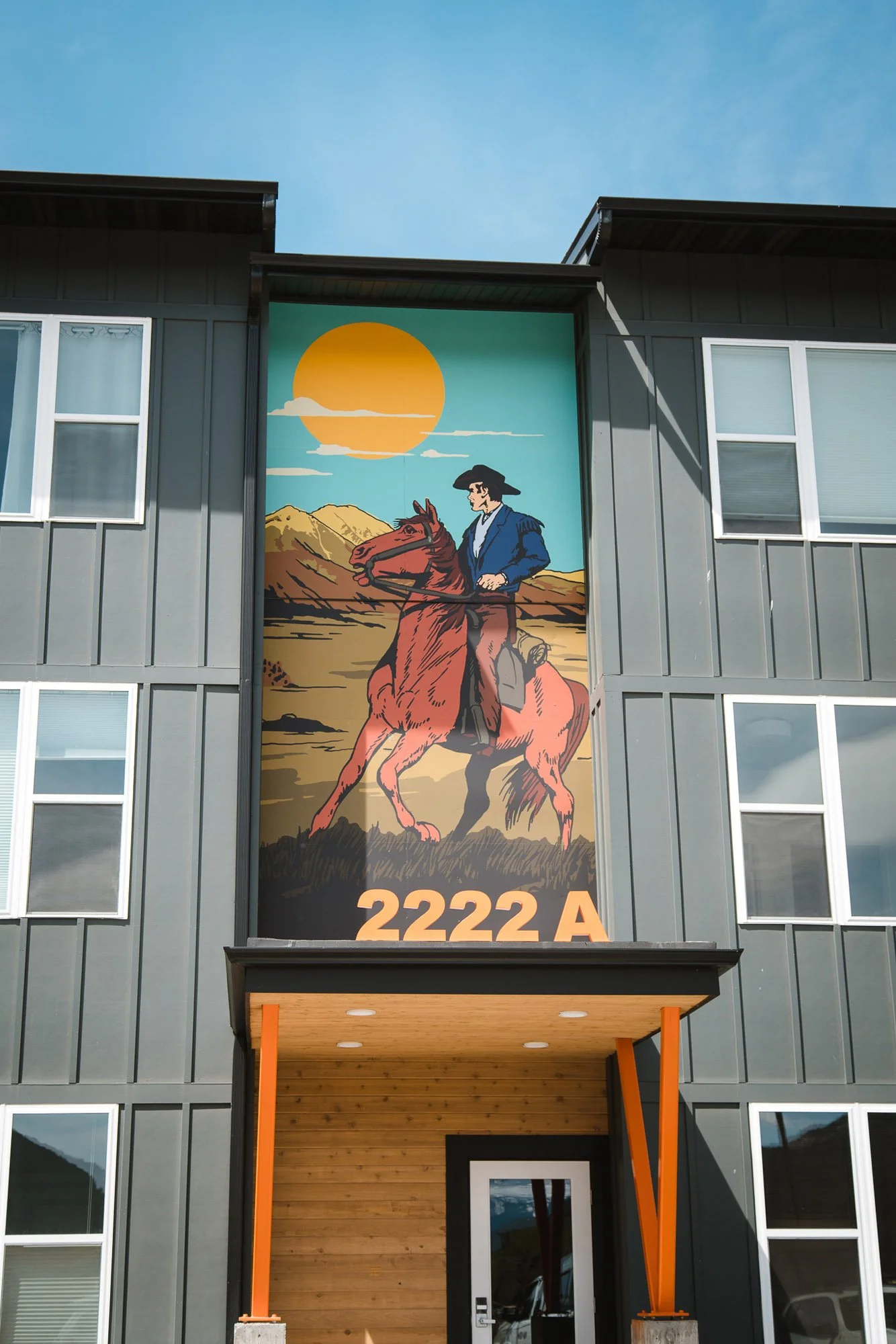 Striking wall wrap above a townhouse doorway showing a cowboy on a red horse riding through a desert landscape with mountains and a setting sun, labeled “2222A” in Bozeman, MT.