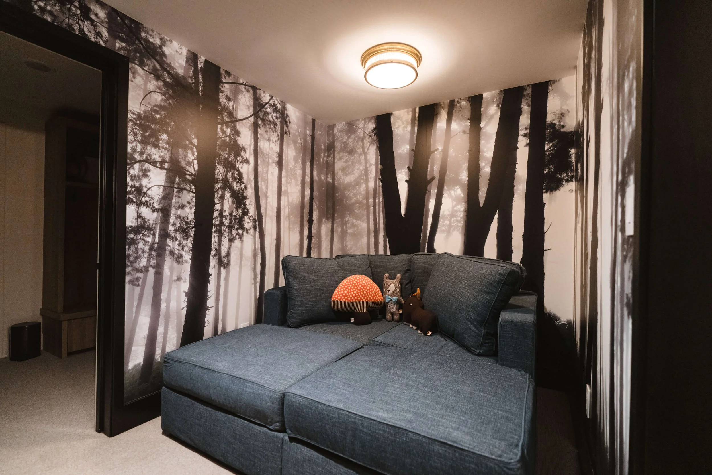 Cozy children's reading nook with a misty forest wall wrap behind a blue sectional sofa strewn with plush toys. Location: Big Sky, MT.