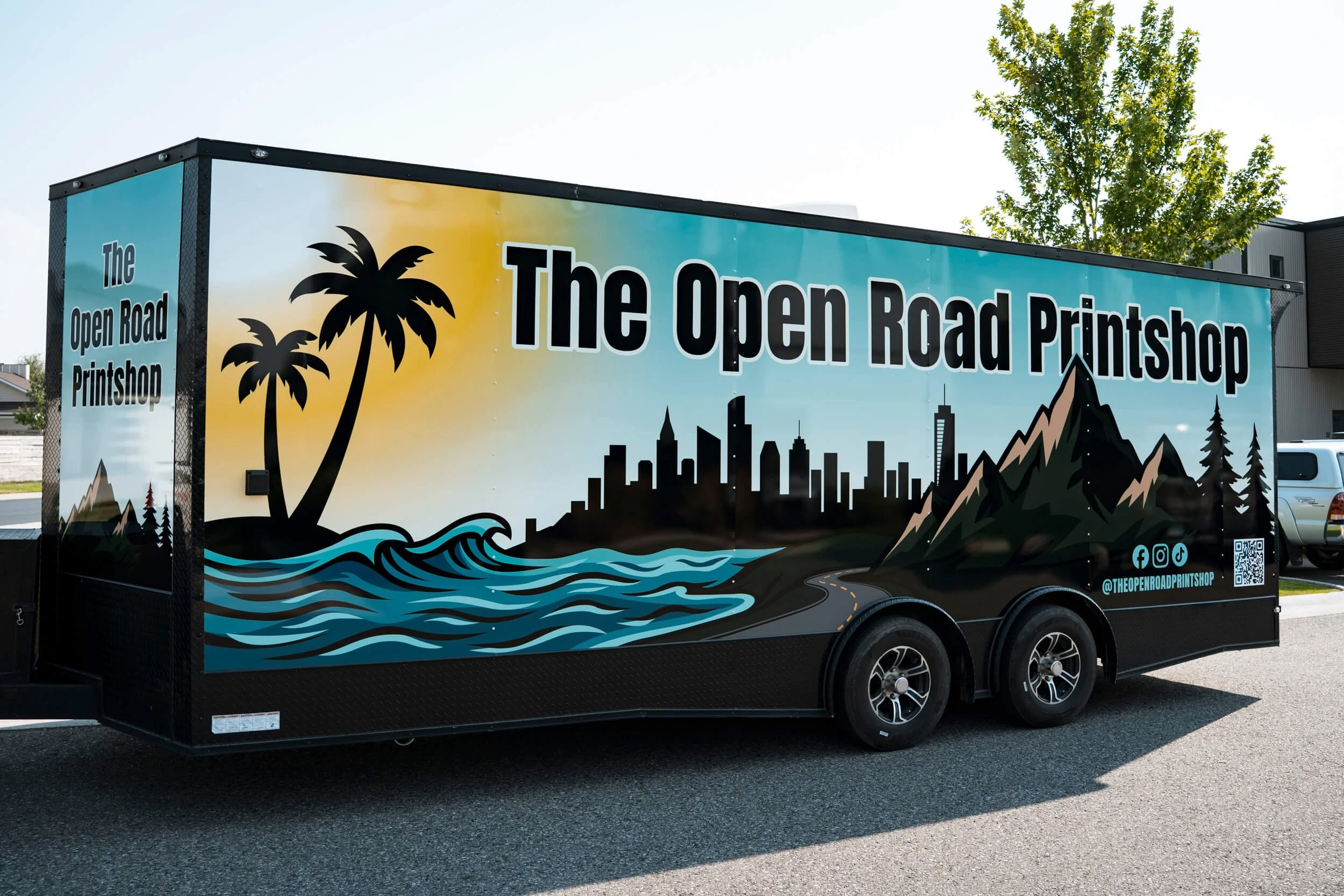 Vinyl trailer wrap with palm trees, the ocean, city skyline, mountains and trees all on a blue background. Large black text runs the length of the trailer for The Open Road Printshop
