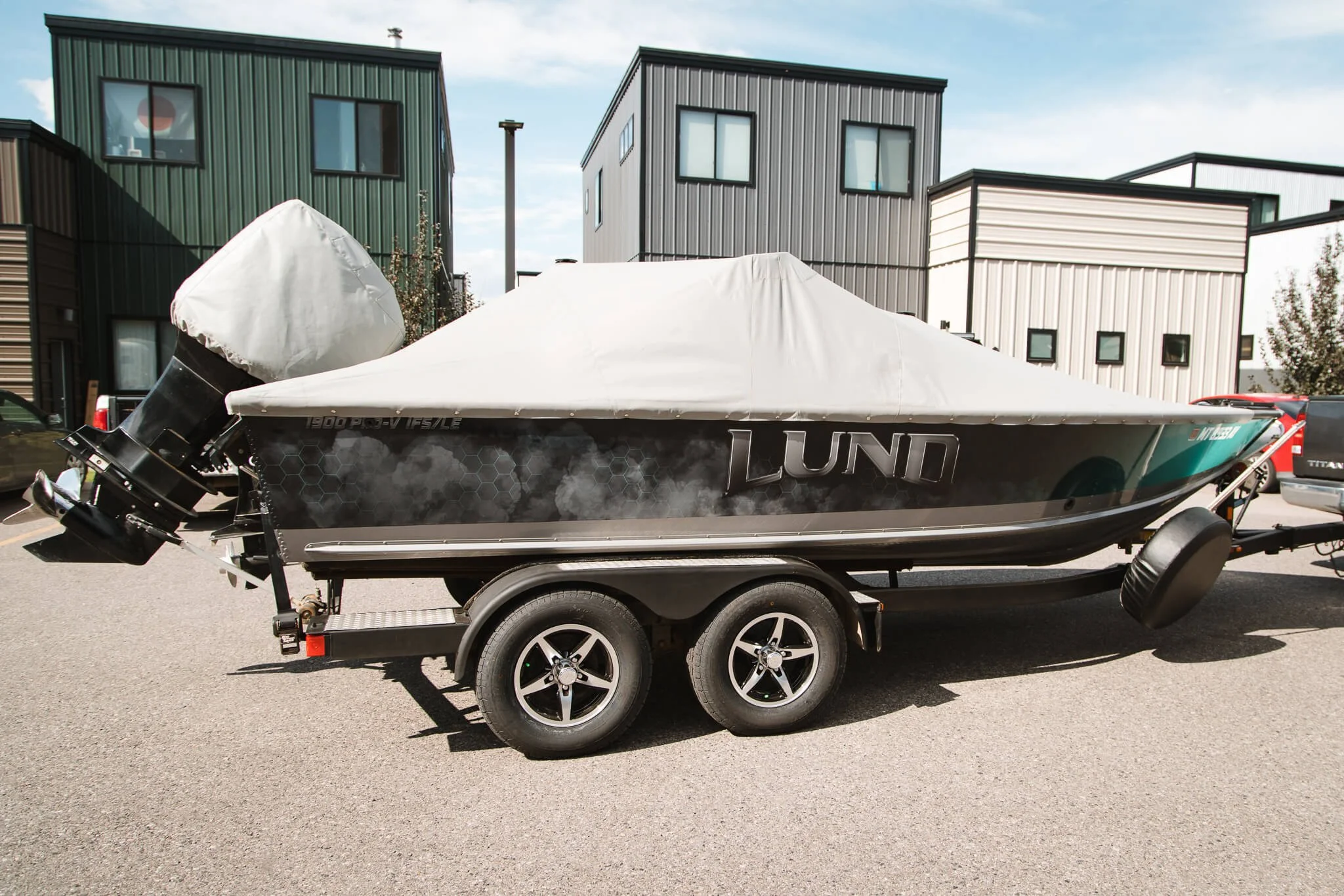 LUND boat featuring a weathered-style gray and teal custom vinyl wrap, partially covered and parked in a modern commercial area in Bozeman, MT.