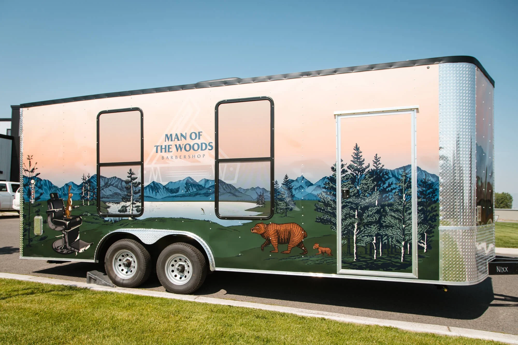 Full-color trailer wrap for Man of the Woods Barbershop in Bozeman, Montana, depicting a lakeside wilderness scene with barbershop chair, bear, and mountain landscape. 