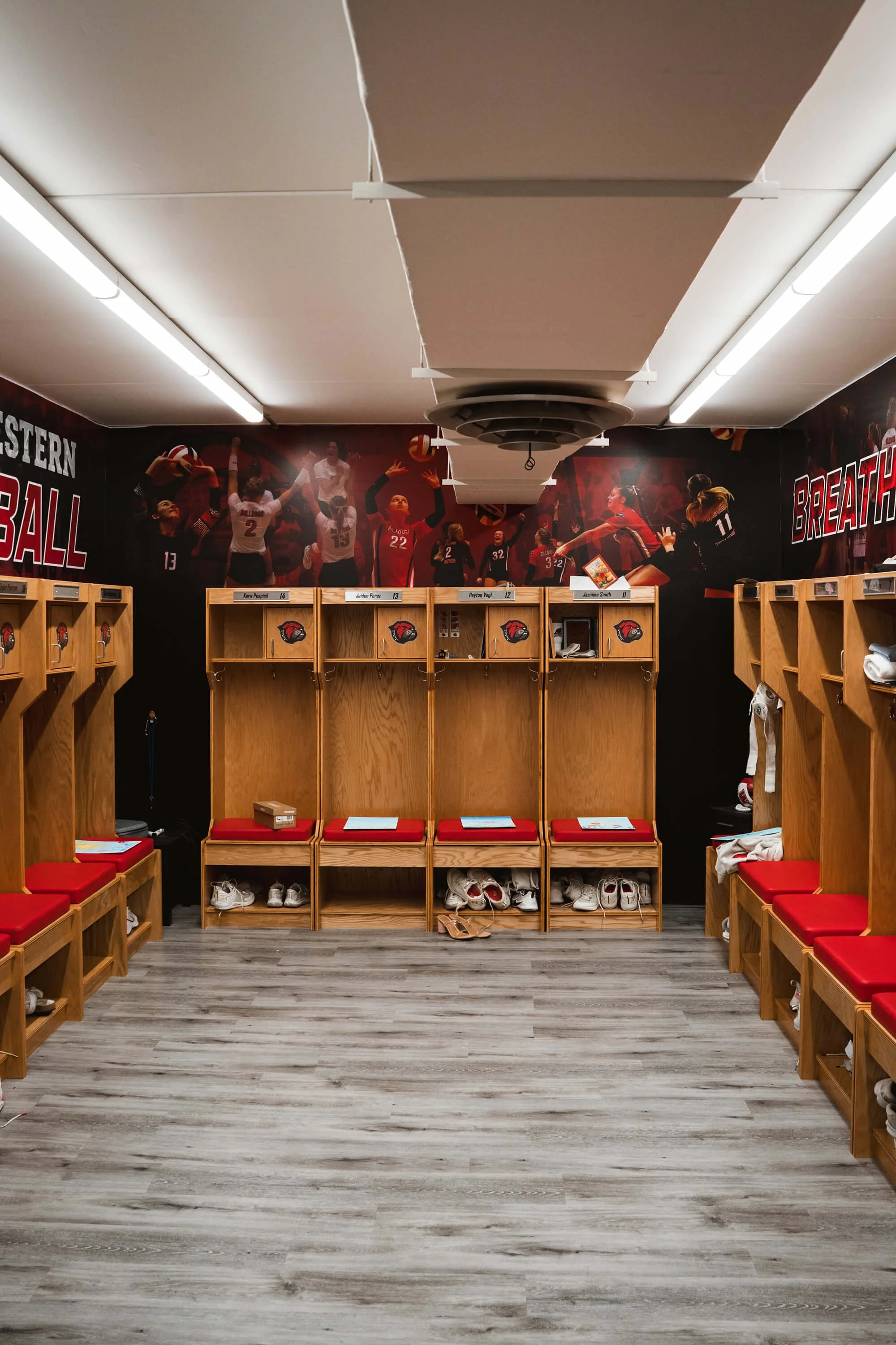 Custom sports locker room wall wrap with volleyball players in action and the word “BREATHE” wrapping around the upper wall above built-in wooden lockers and red bench cushions. Business Location: UM Western in Dillon, MT.