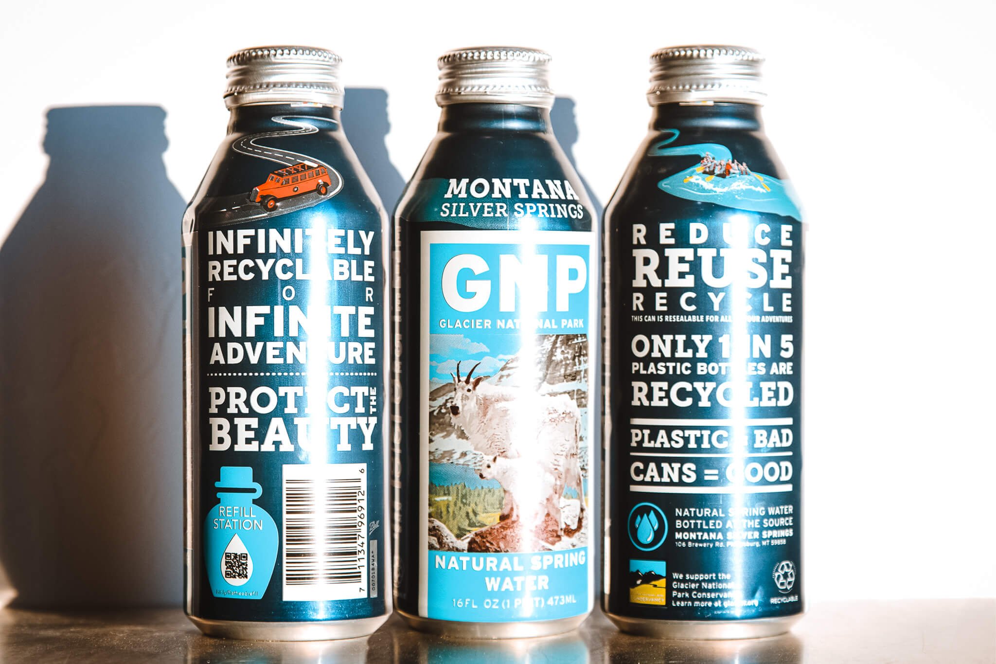 Three metal water bottles with recycling messages and Glacier National Park imagery, emphasizing sustainability and natural spring water from Montana.