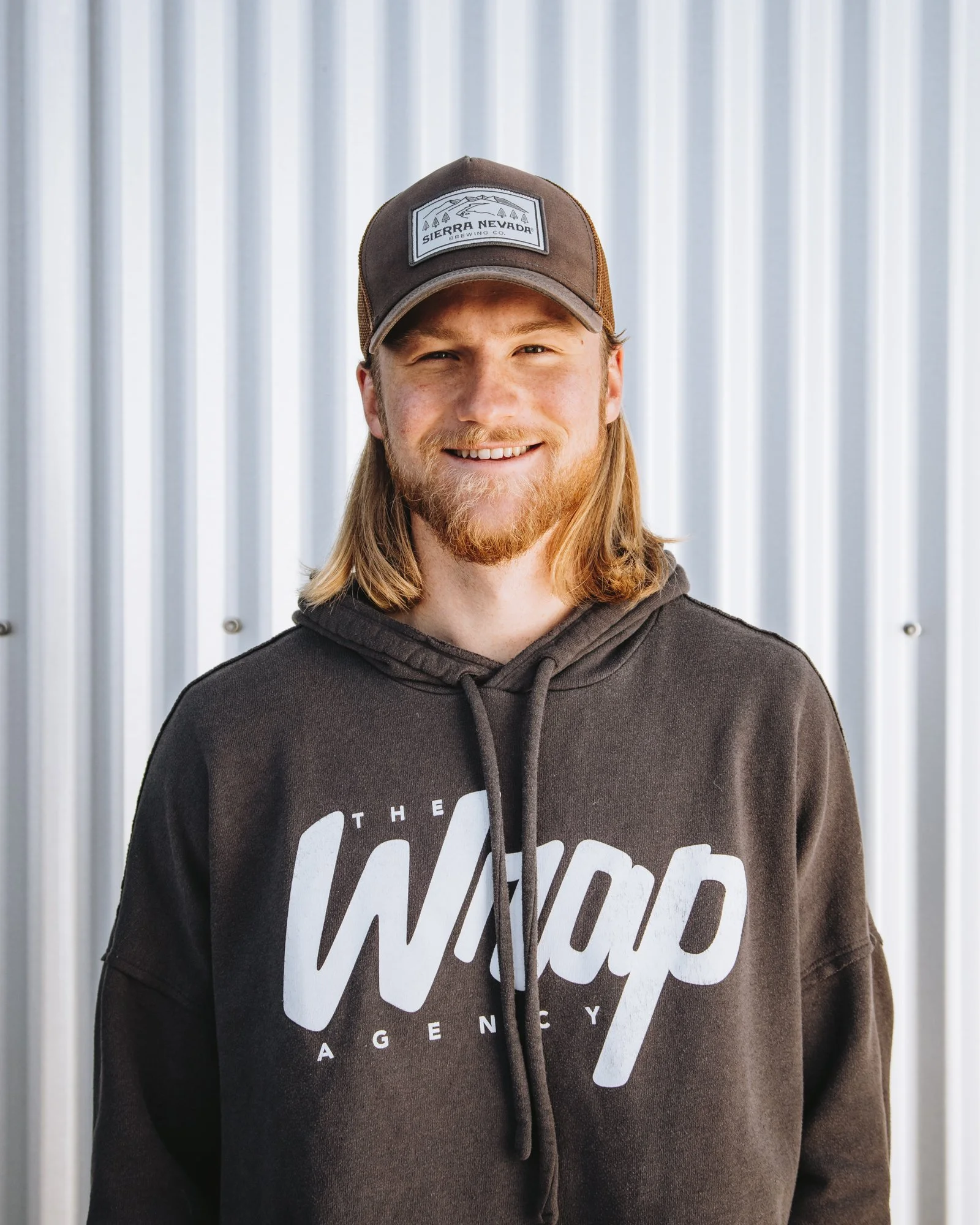 Person wearing a brown cap and black hoodie with "The Wrap Agency" logo, standing in front of a corrugated metal wall.