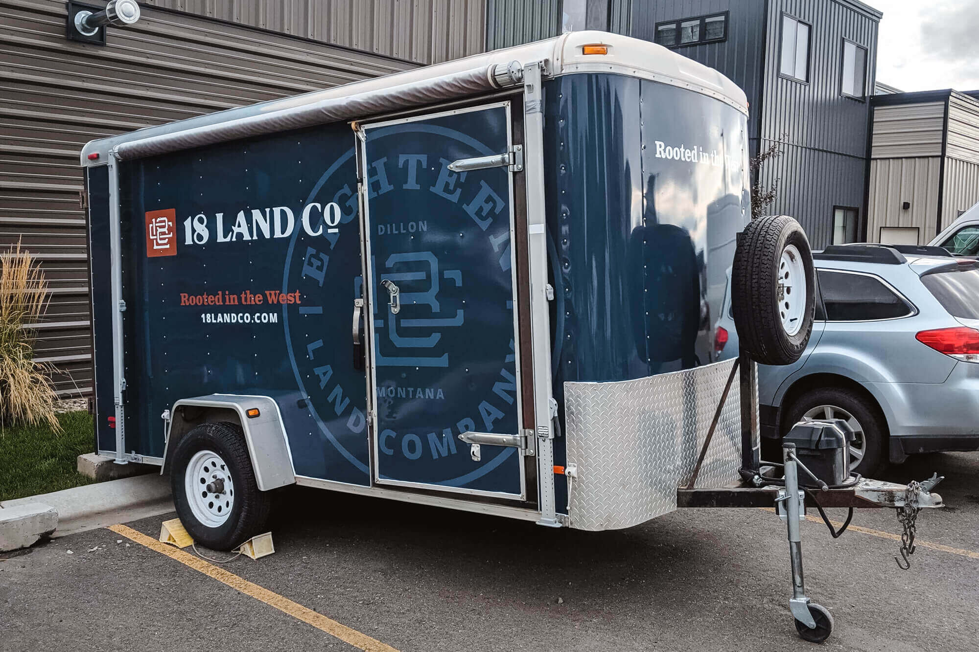 Side view of a branded trailer for 18 Land Co in Bozeman, Montana, with custom trailer graphics including the company logo, slogan “Rooted in the West,” and a bold navy blue background.