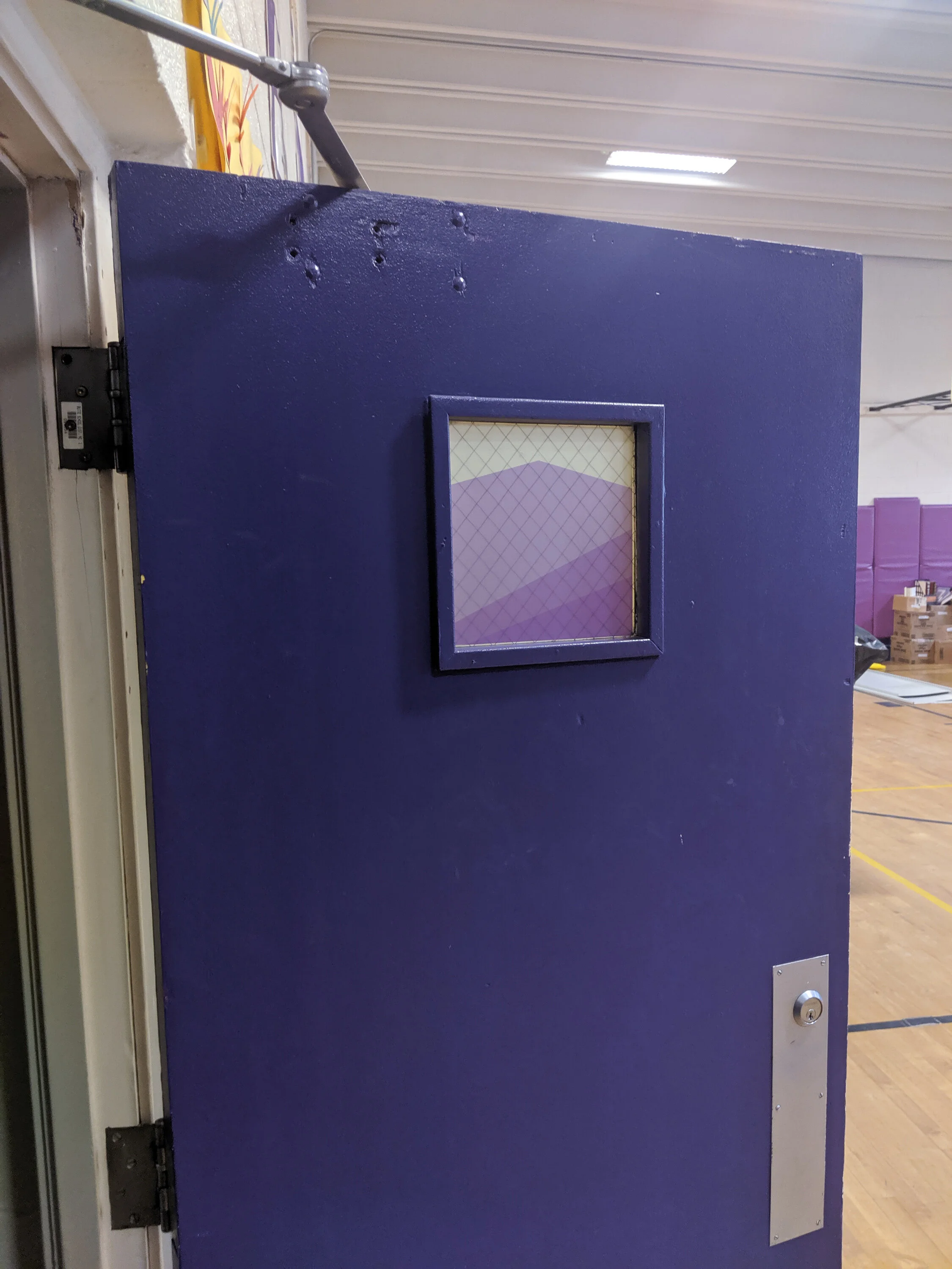 Purple gym door with a small square window with frosted vinyl, shown slightly open, revealing a gymnasium interior with wooden floors and sports equipment in the background.