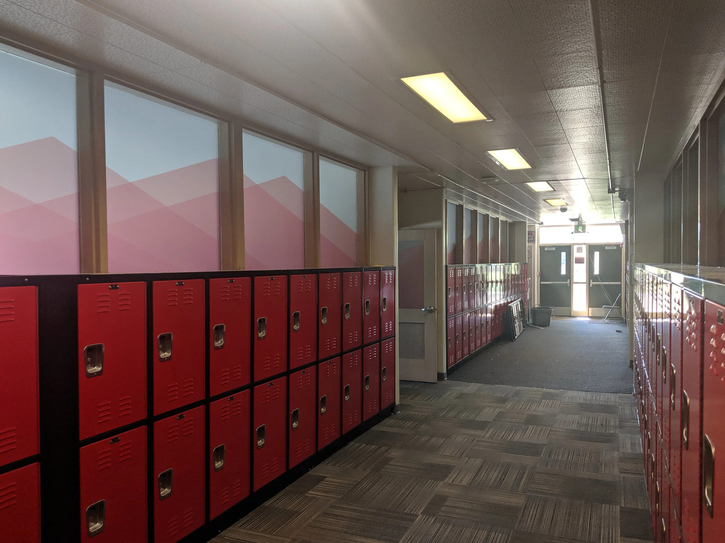 School hallway with red lockers and patterned carpet flooring. Windows above lockers have mountain scenes printed on frosted privacy film. In Butte, Montana.