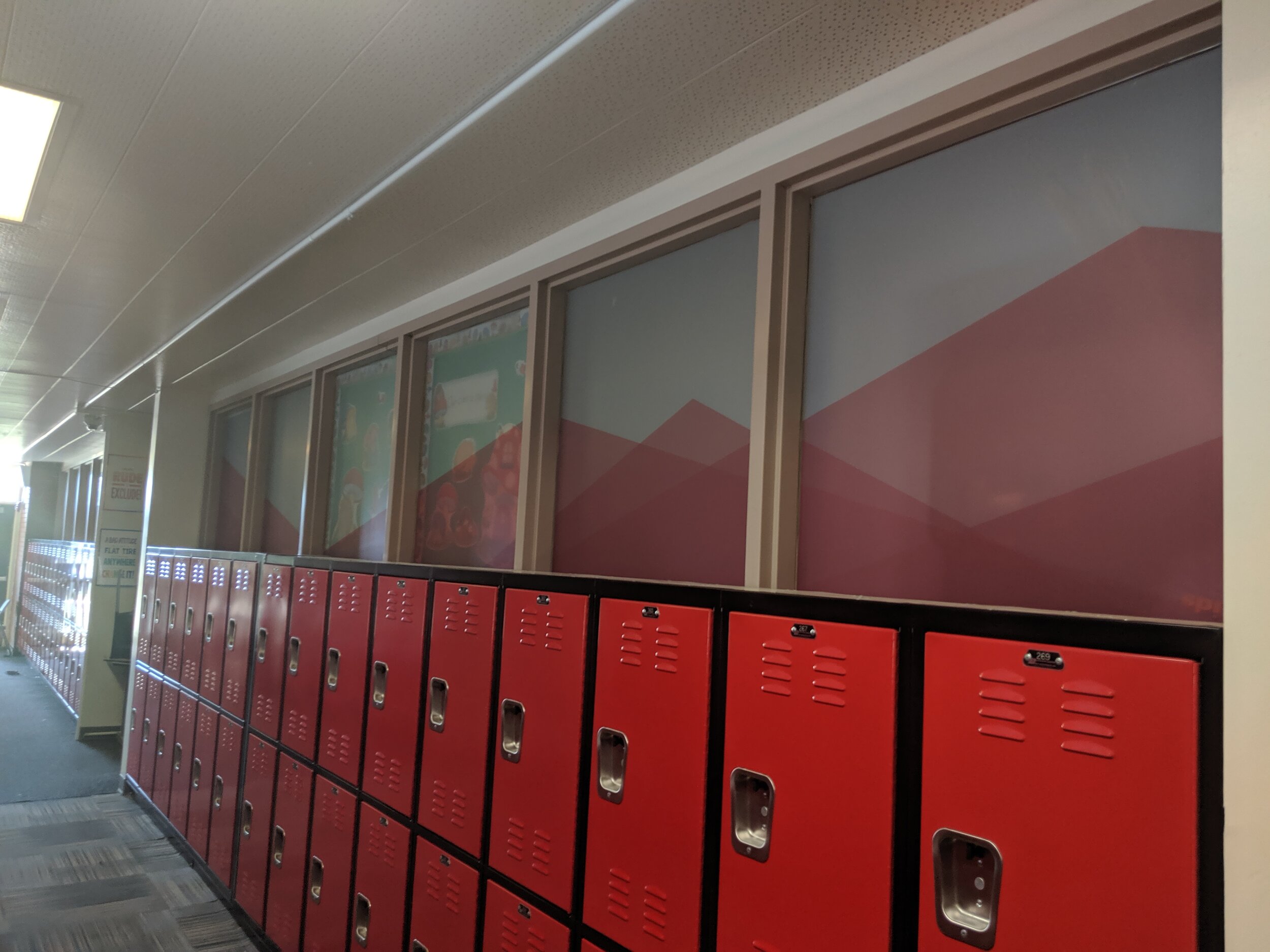 Red lockers in a school hallway with windows above them, featuring a geometric printed design on frosted privacy film and a ceiling with fluorescent lighting. In Butte, Montana.