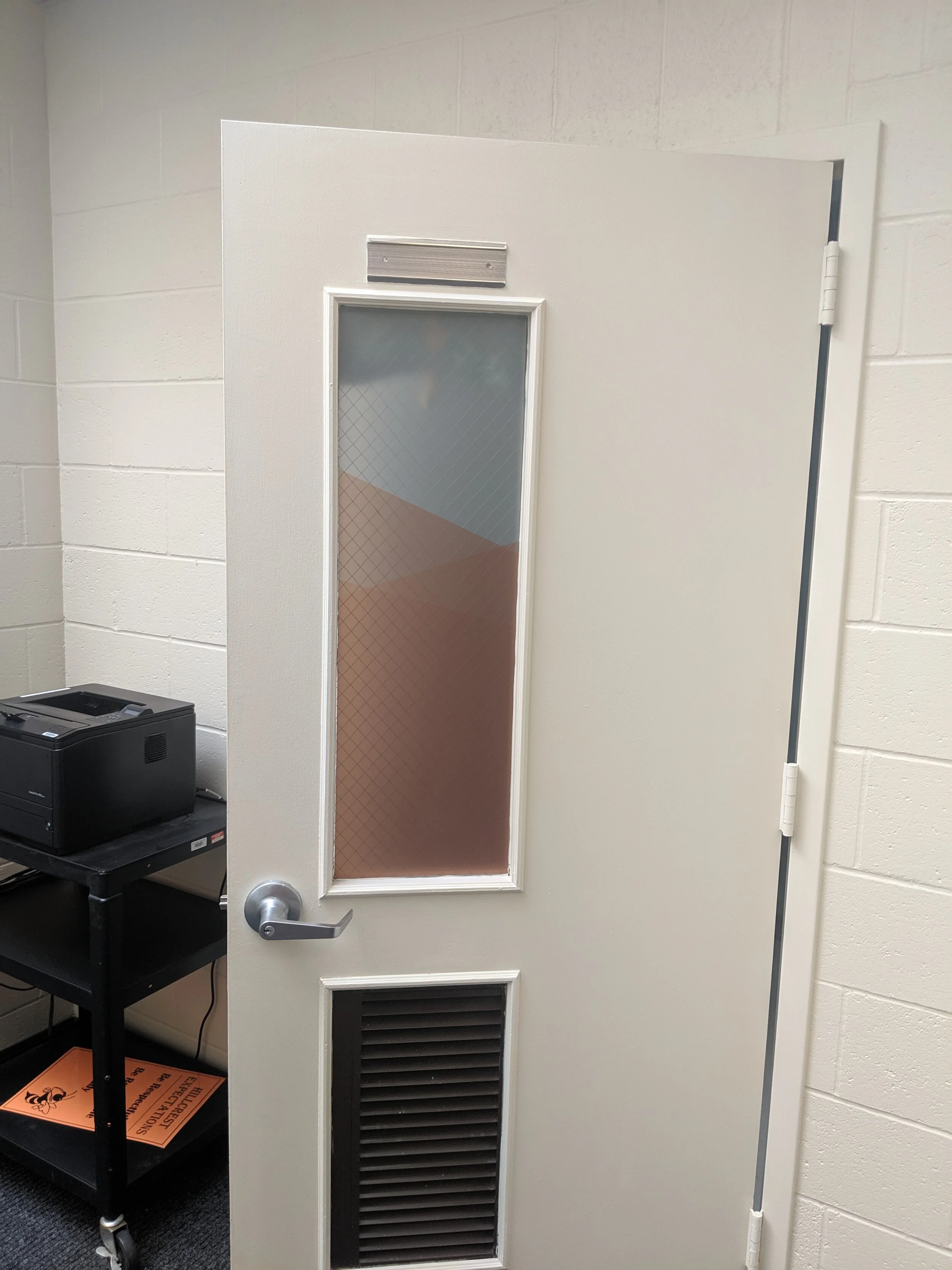 Open door with frosted window and ventilation grille, inside a room with a printer on a cart and a white brick wall background. In Butte, Montana.