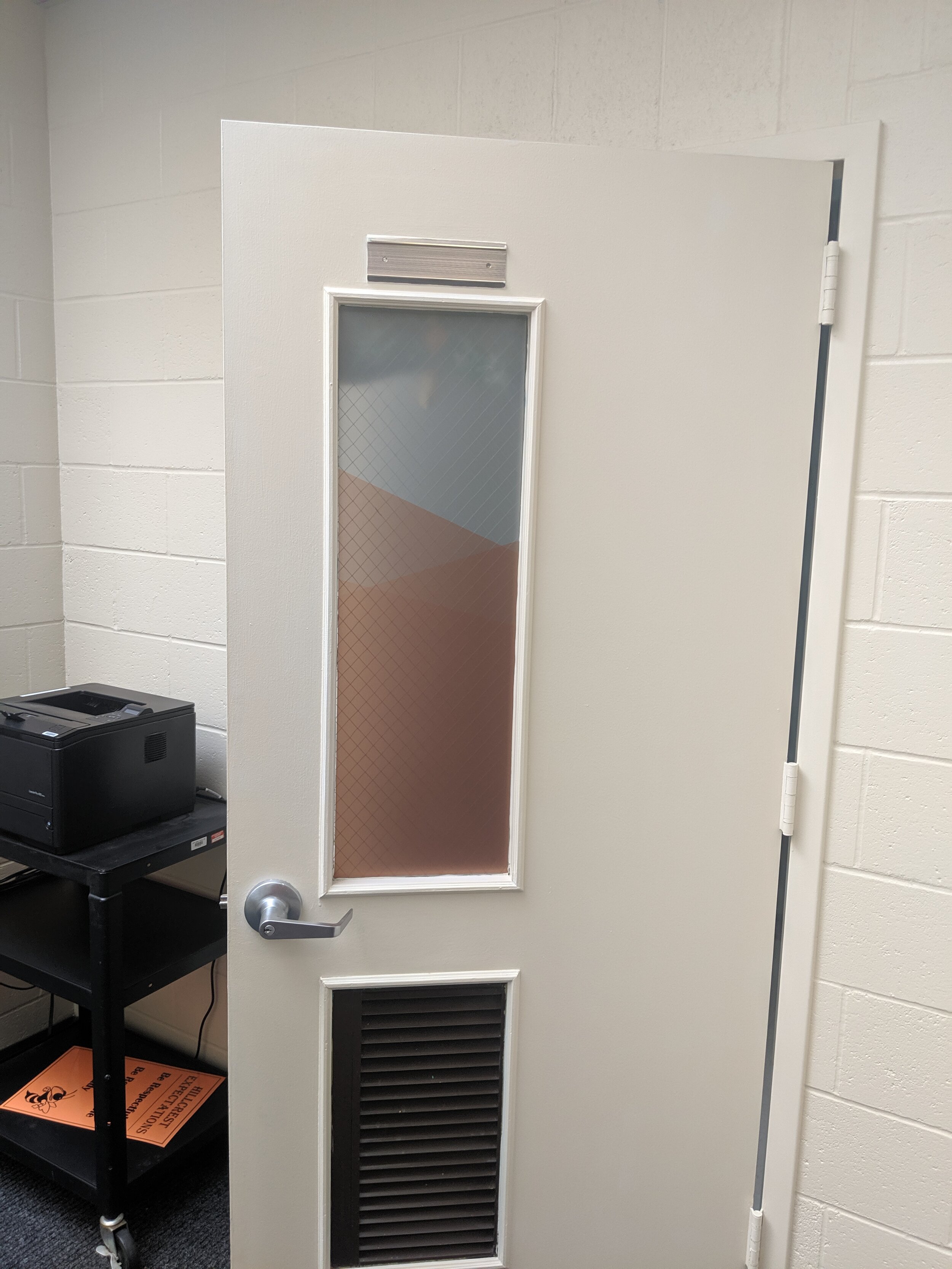 Open door with frosted window and ventilation grille, inside a room with a printer on a cart and a white brick wall background. In Butte, Montana.