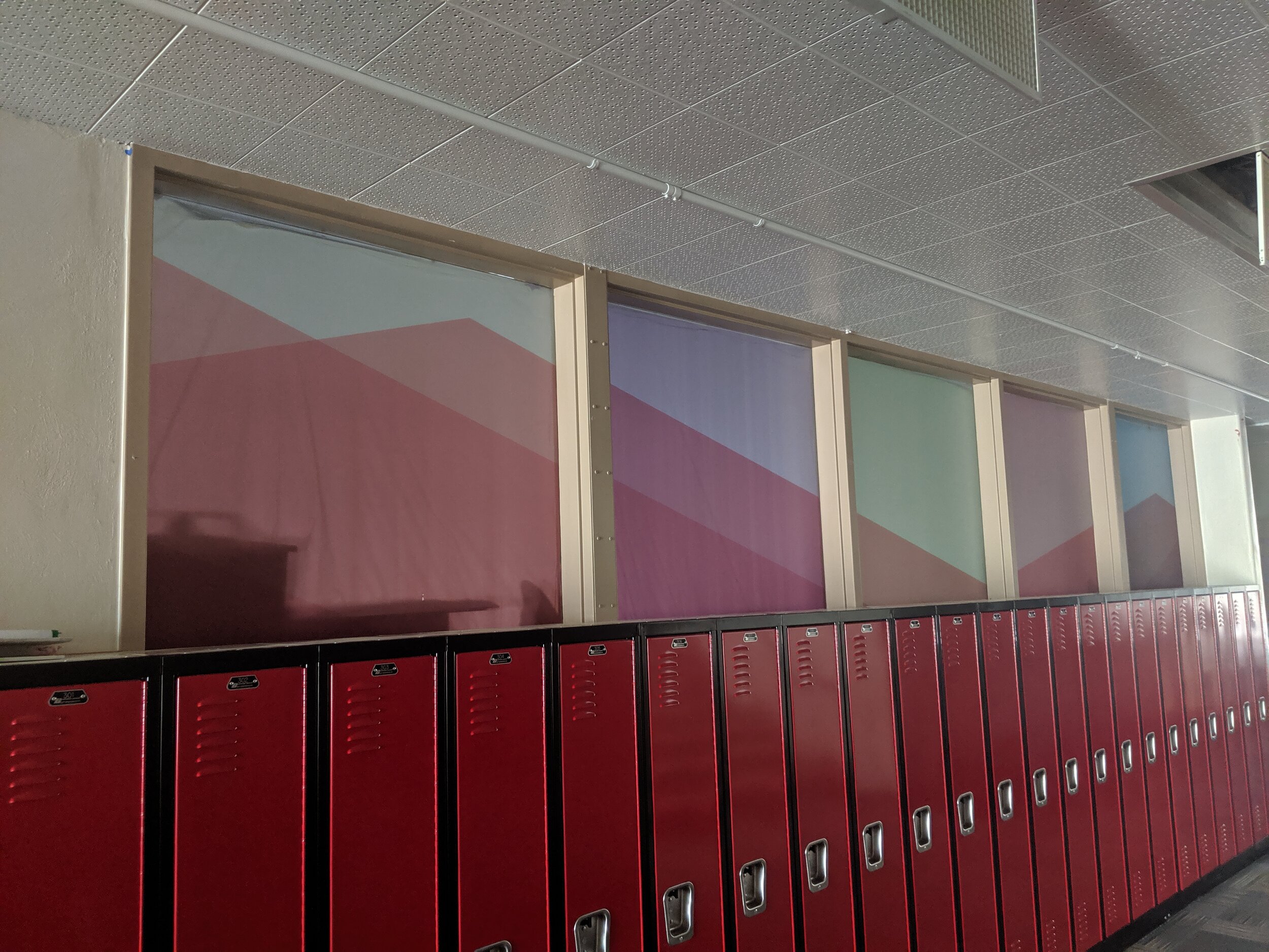 Red lockers lined against a wall beneath windows with mountain scenes printed on frosted privacy film in a school hallway.