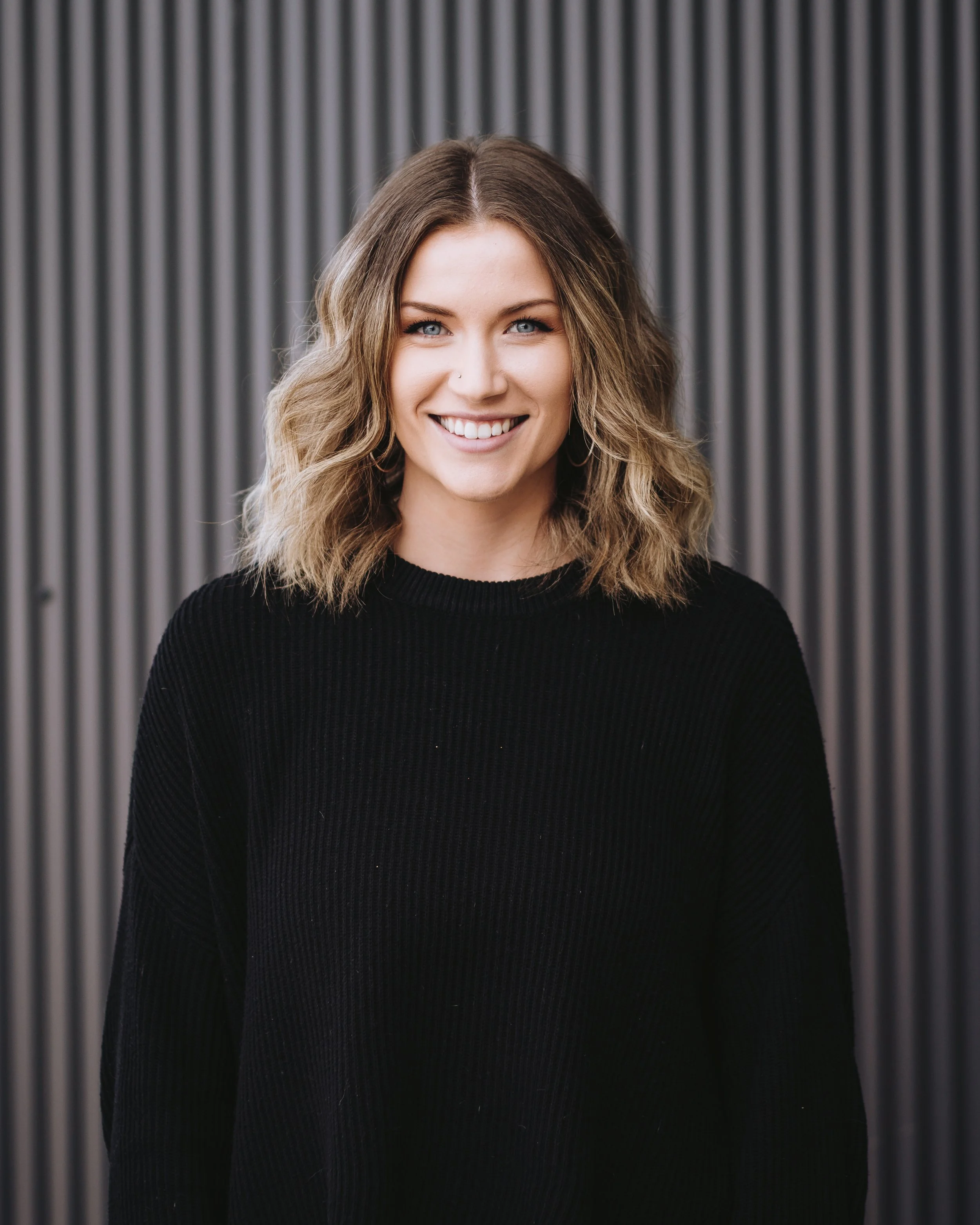 Smiling person with wavy hair wearing a black sweater in front of a corrugated metal background.