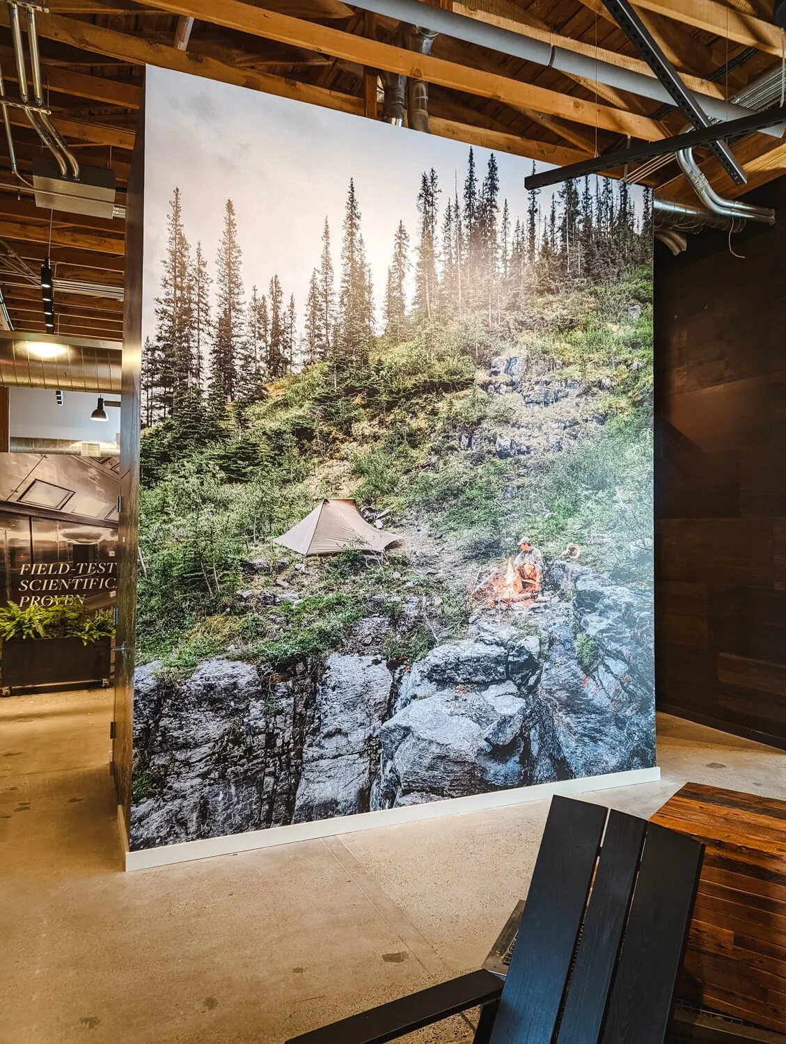 Dramatic wall wrap showcasing a tent and campfire perched on a rocky cliffside surrounded by evergreen forest, featured in a high-ceiling commercial interior at Sitka Depot in Bozeman, MT.