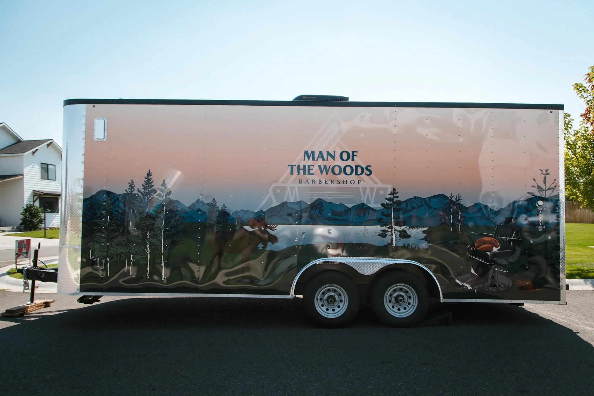 Custom trailer graphics for Man of the Woods Barbershop in Bozeman, Montana, featuring a scenic forest and mountain landscape with a moose, lake, and barber chair illustration on a mobile trailer wrap.