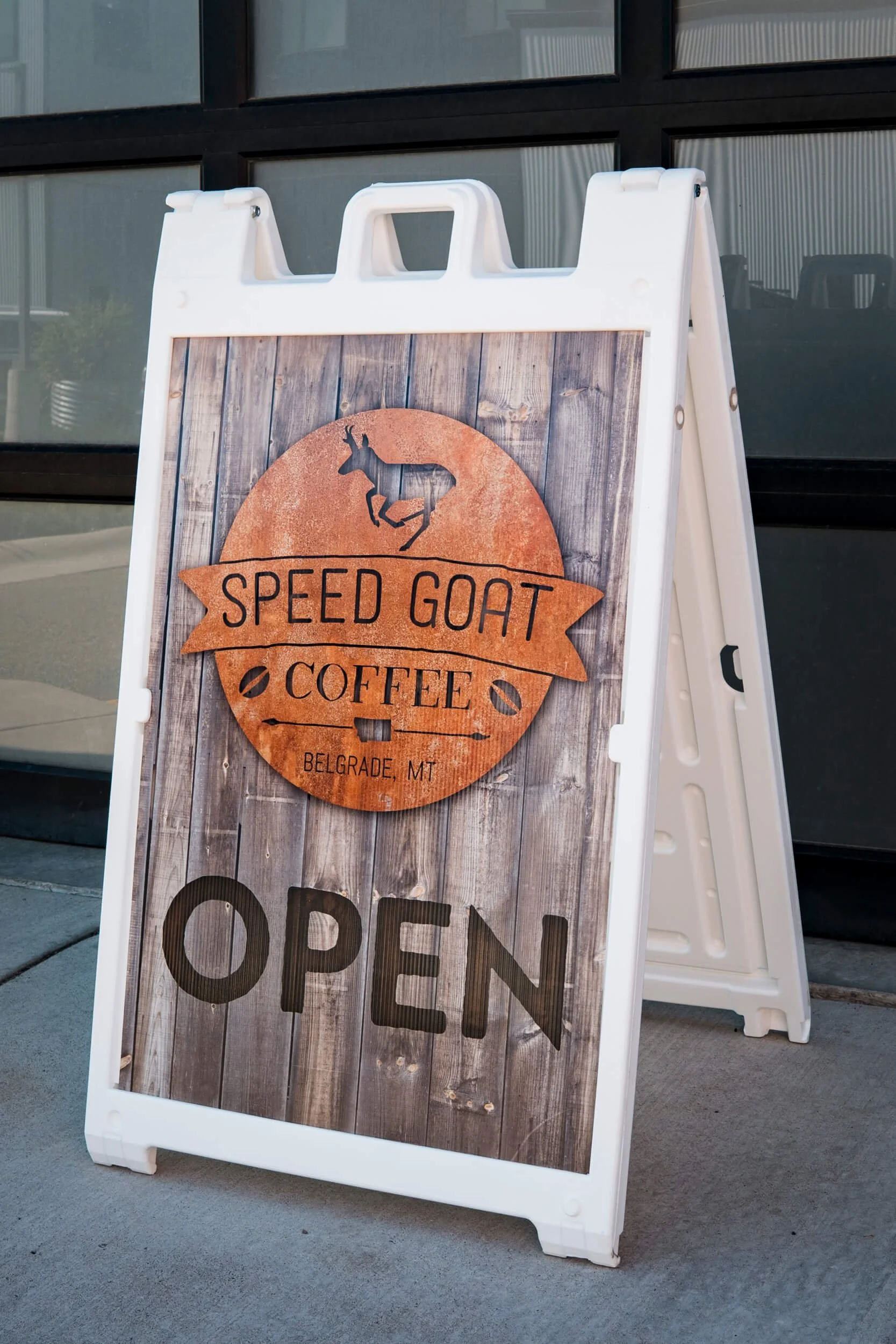 White sandwich board frame with a coroplast sign insert with a rusty metal Speed Goat Coffee graphic with the word "Open" below, all on a barnwood background